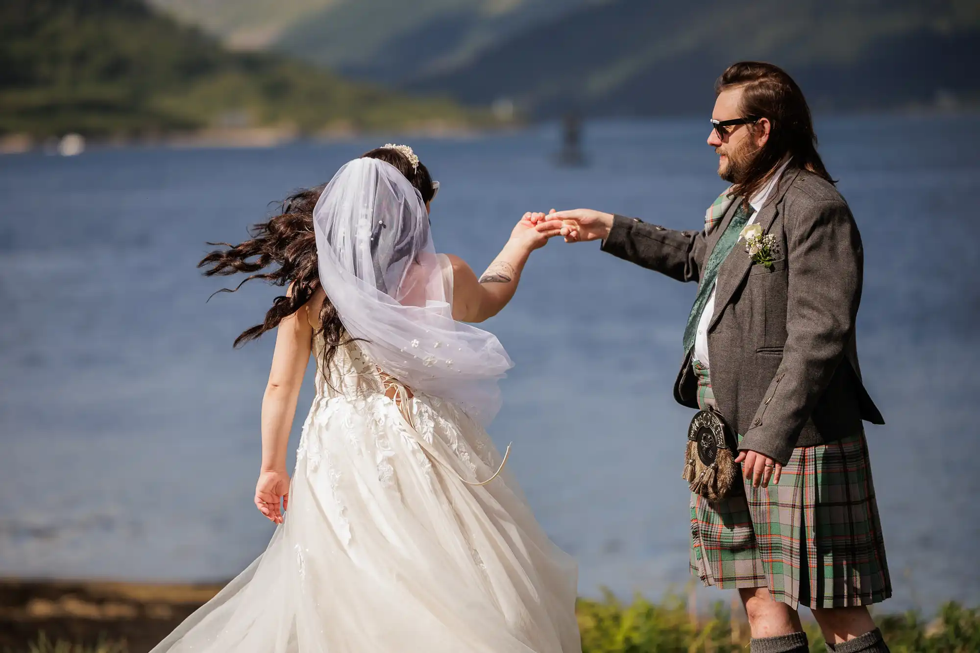 A bride in a white dress and veil holds hands with a groom wearing a grey jacket and traditional tartan kilt. They stand outdoors near a blue lake, with green hills and mountains in the distant background, enjoying a sunny day.
