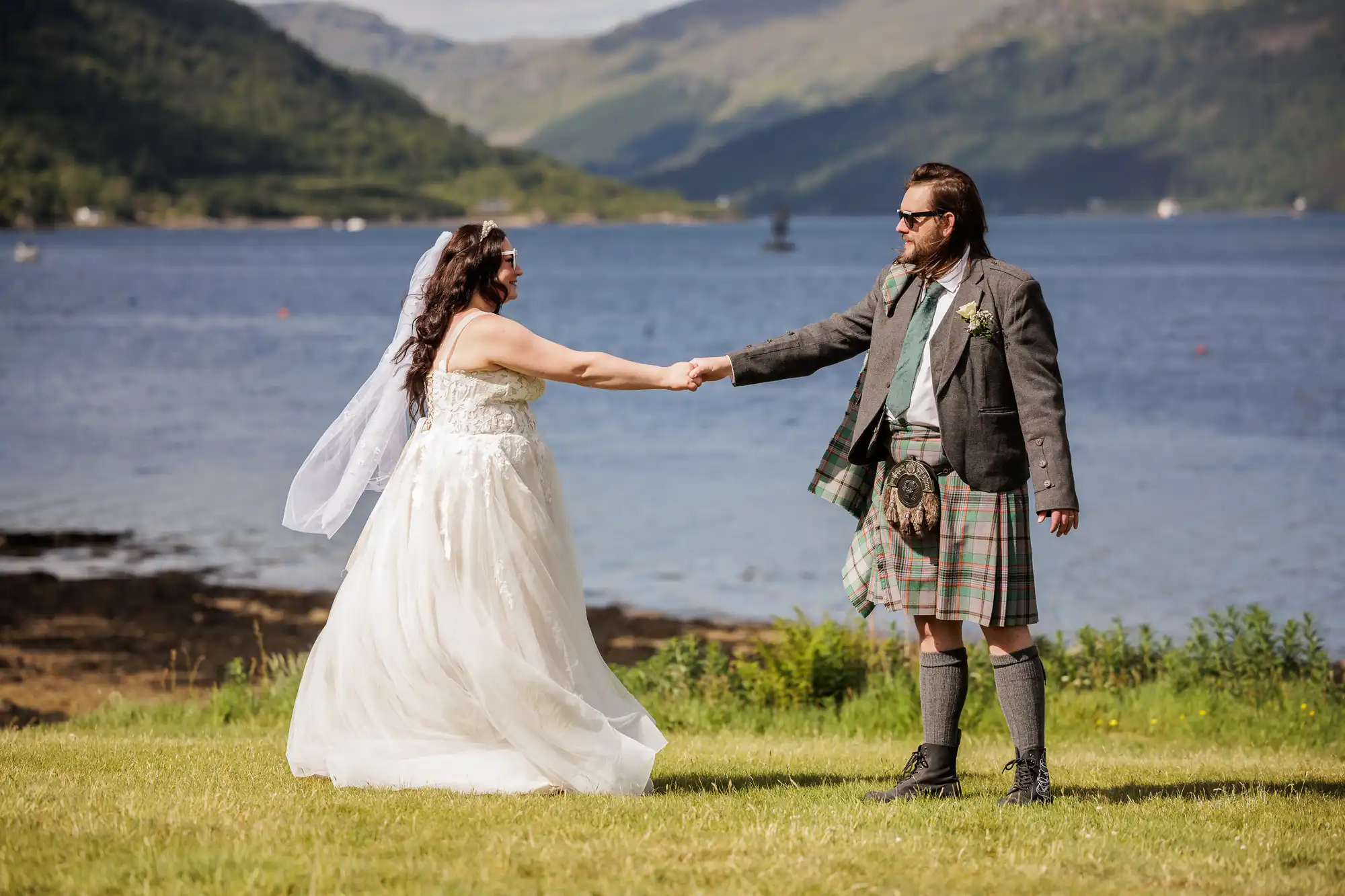 A bride in a white dress and veil holds hands with a groom in a kilt and suit jacket. They stand on green grass by a scenic lake, with green mountains and a blue sky in the background, sharing a joyful moment outdoors.
