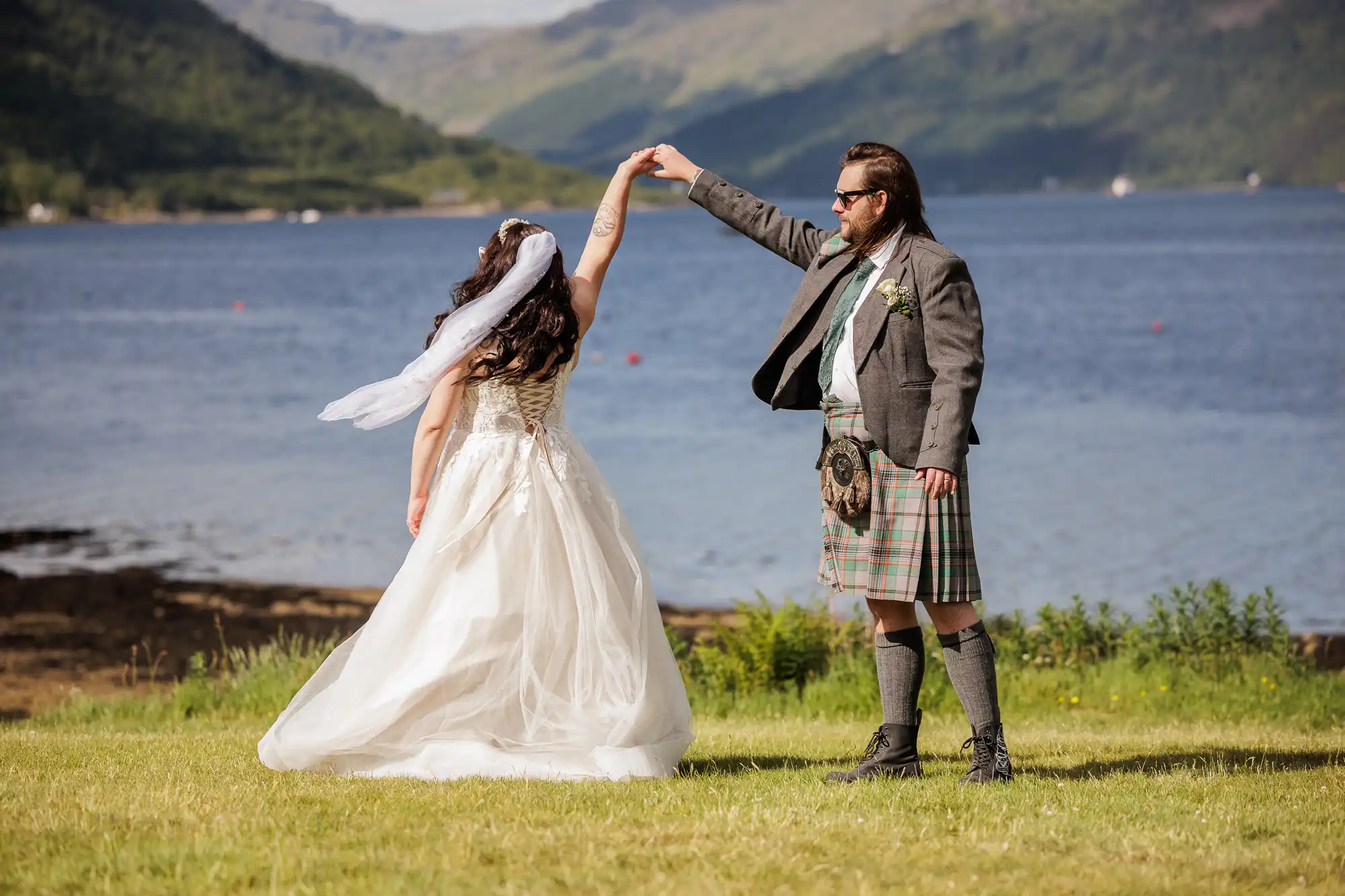 A bride in a white gown and veil spins under the raised hand of a groom in a grey jacket, kilt, and sporran. They stand on grass beside a lake, with green hills and mountains in the background under a clear sky.