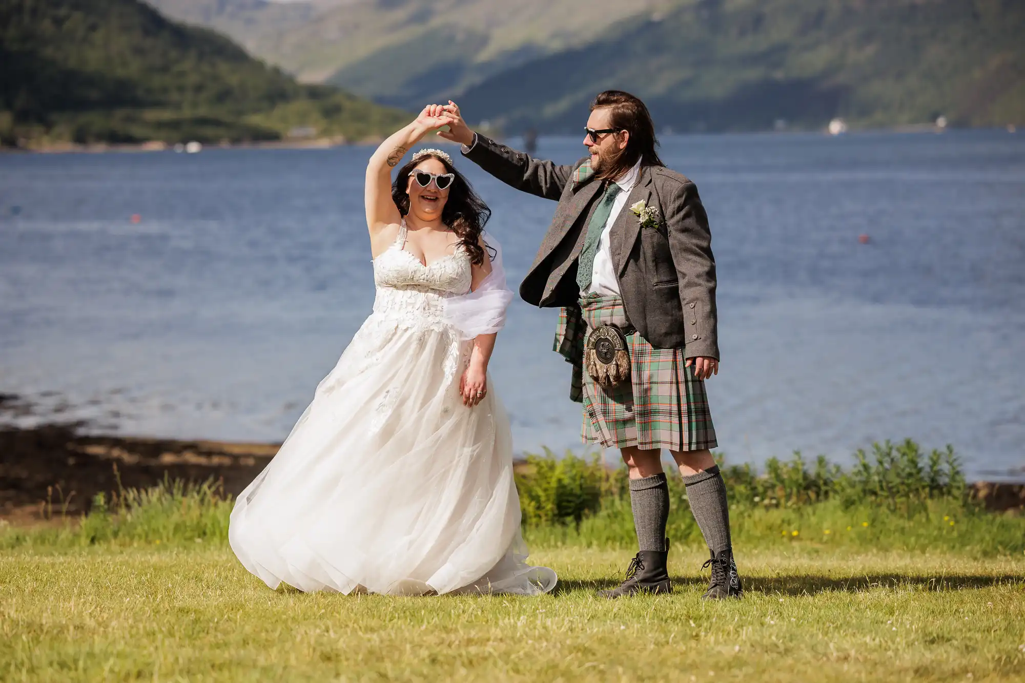 A bride in a white dress and sunglasses twirls with a groom wearing a grey jacket, kilt, and sunglasses, on grass by a lake with mountains and blue sky in the background; they both smile and appear joyful.