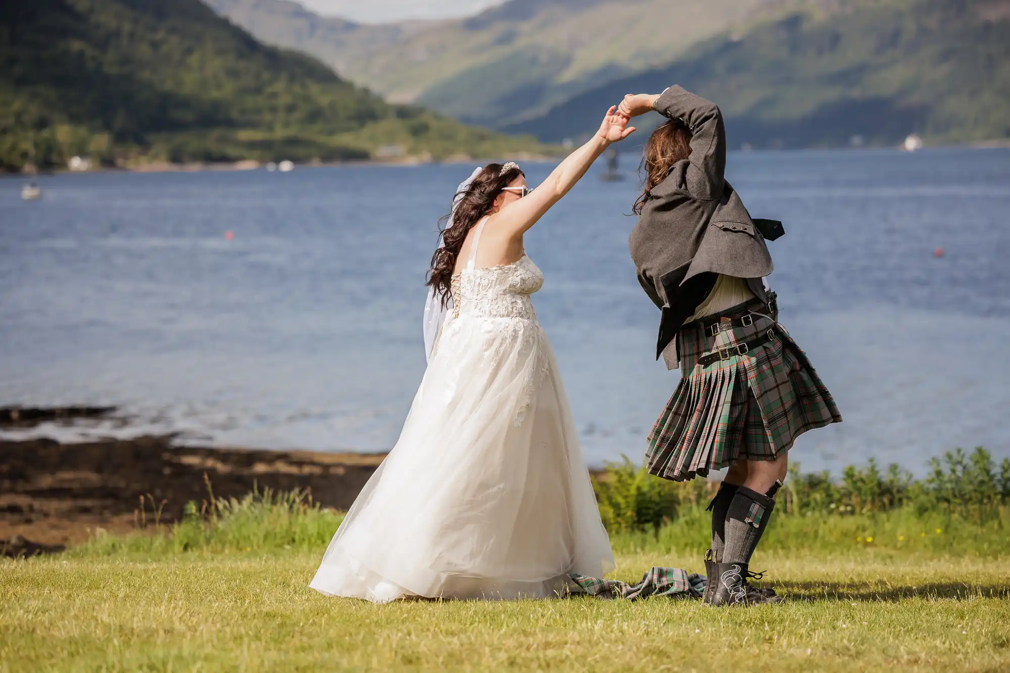 A bride in a white dress and a groom in a kilt dance joyfully on grass near a lake, with rolling, green hills and blue mountains in the background under a clear sky. The playful couple hold hands, celebrating outdoors.