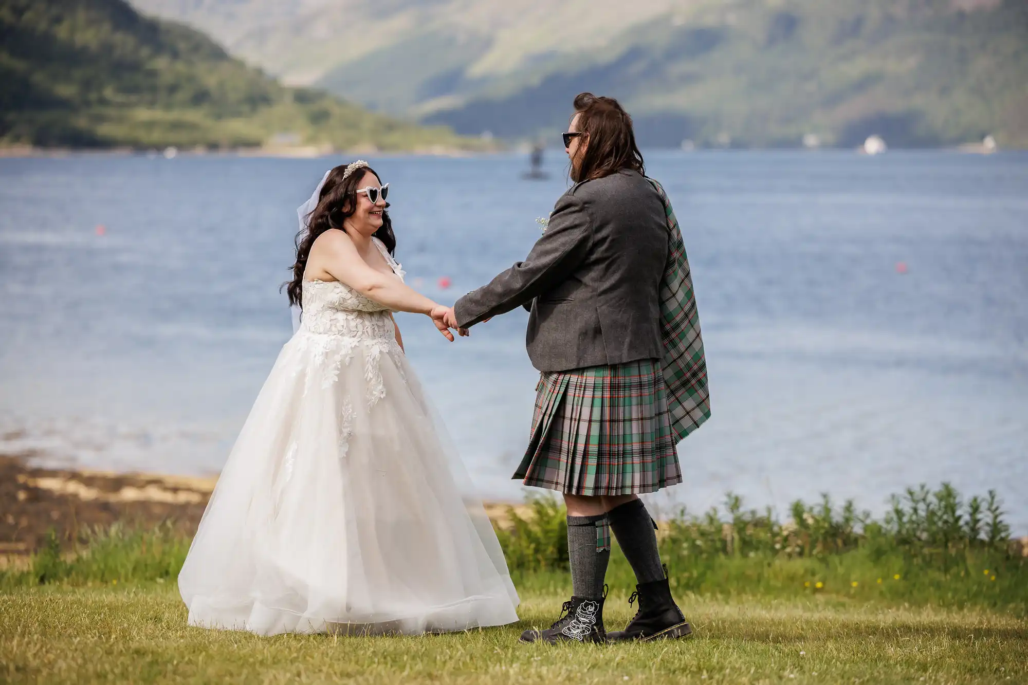 A couple stands on grass by a lake with green hills in the background. One wears a white wedding dress and sunglasses; the other wears a grey jacket, checked kilt, and boots. They hold hands, smiling at each other, enjoying a sunny outdoor moment.
