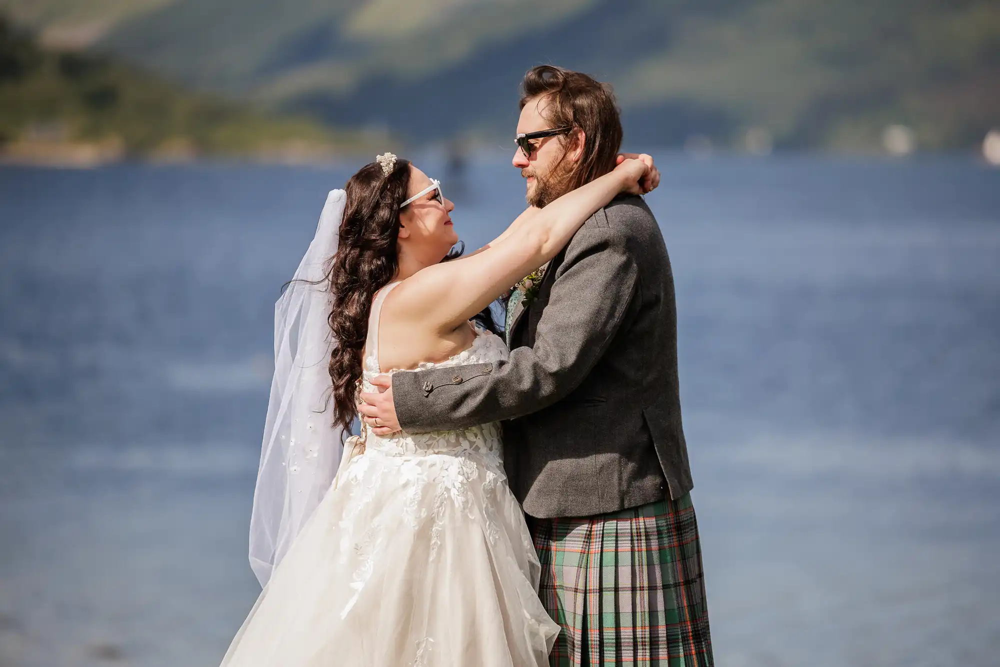 A bride in a white dress and veil embraces a groom in a grey jacket and tartan kilt by a lake, with green hills in the background. They gaze happily at each other under bright sunlight.