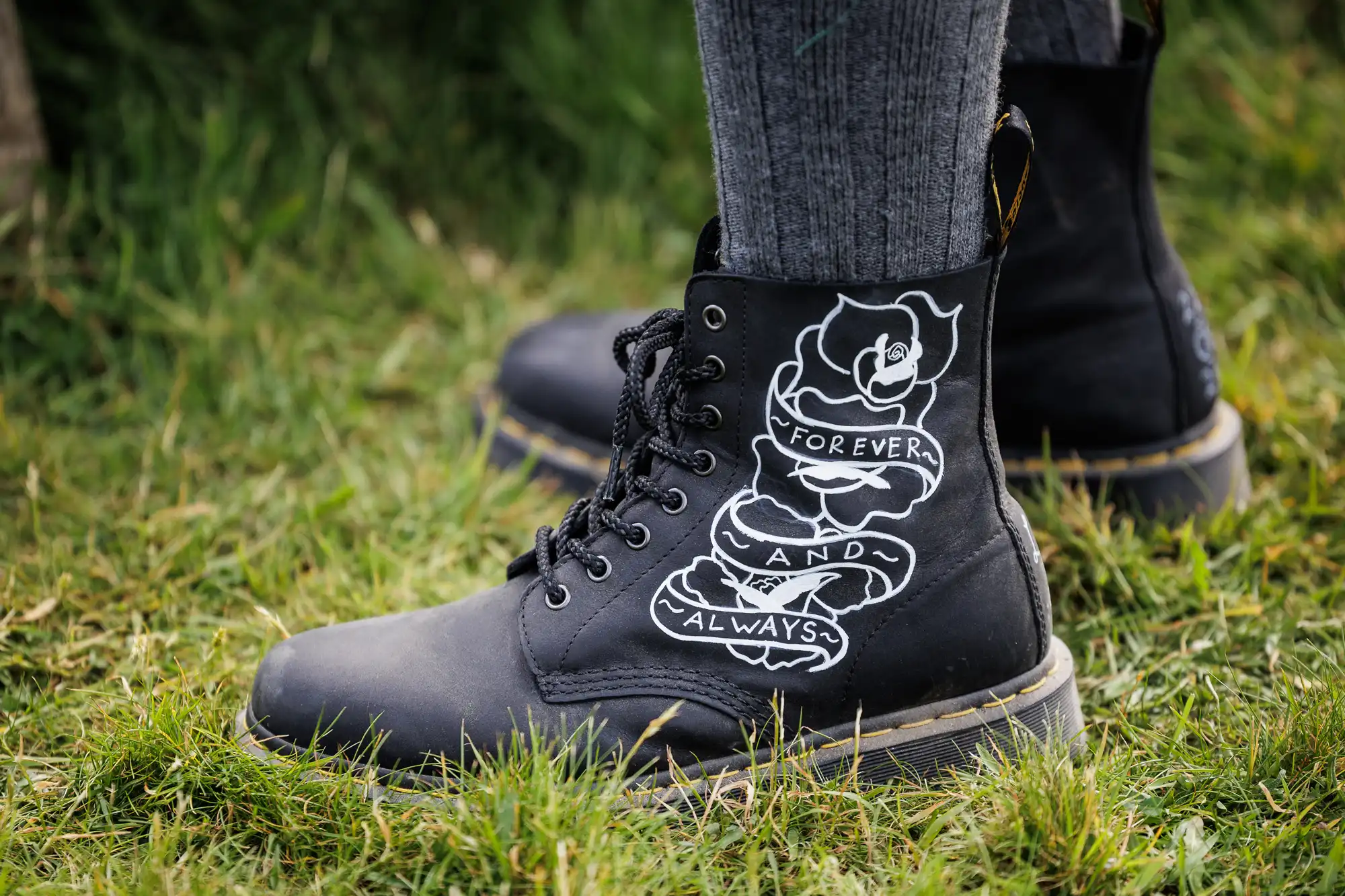 Black lace-up boots with a white rose and a banner illustration on the side, displaying the words “Forever and Always.” The boots are worn with grey socks, and the person is standing on green grass outdoors.
