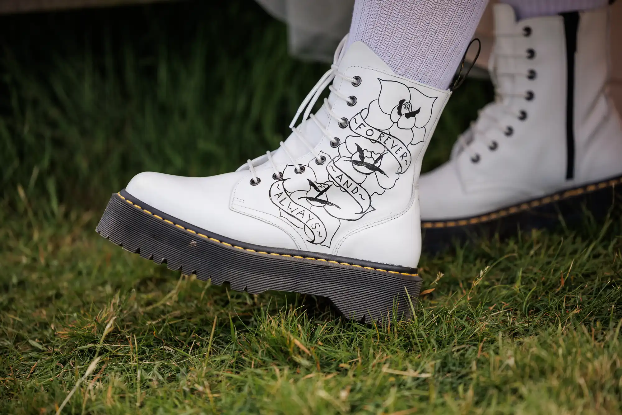 A close-up of a white combat boot with black laces and thick black sole. The side features a tattoo-style design of roses and banners reading “FOREVER,” “AND,” and “ALWAYS.” The boot is worn with white ribbed socks, and is standing on green grass.