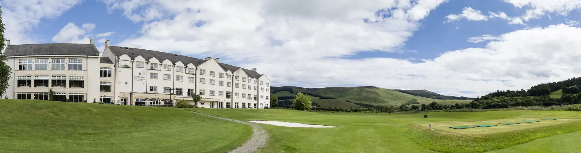 A large white hotel with many windows sits on well-maintained green lawns, bordered by sand bunkers and a golf course. Rolling hills and scattered trees stretch into the background under a partly cloudy blue sky.