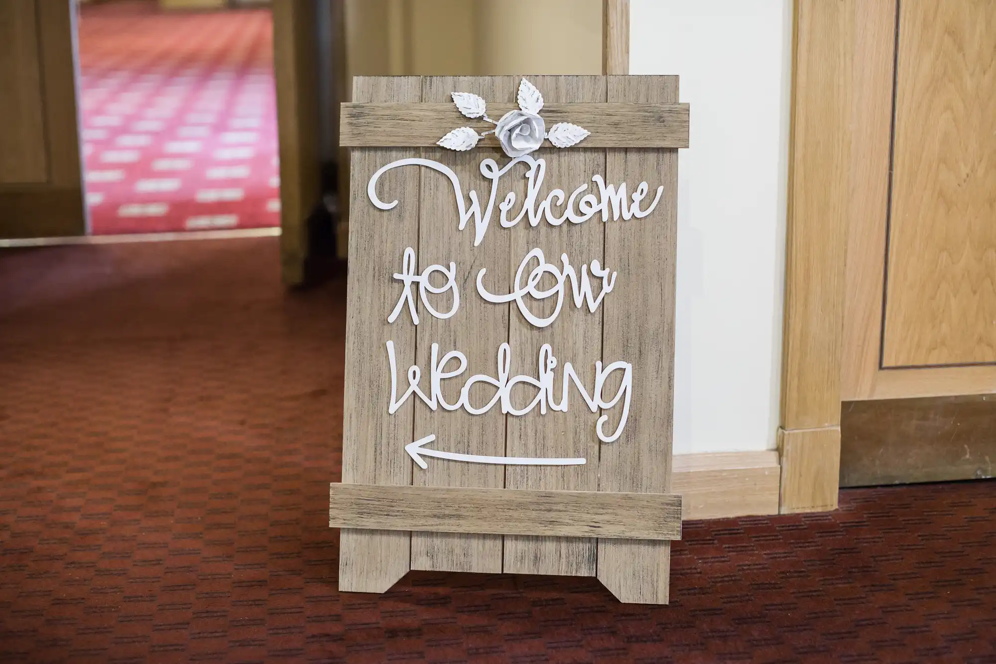 A wooden sign with white decorative lettering and a flower at the top reads, “Welcome to our wedding” with an arrow pointing left. The sign stands on a red-patterned carpet near a doorway.