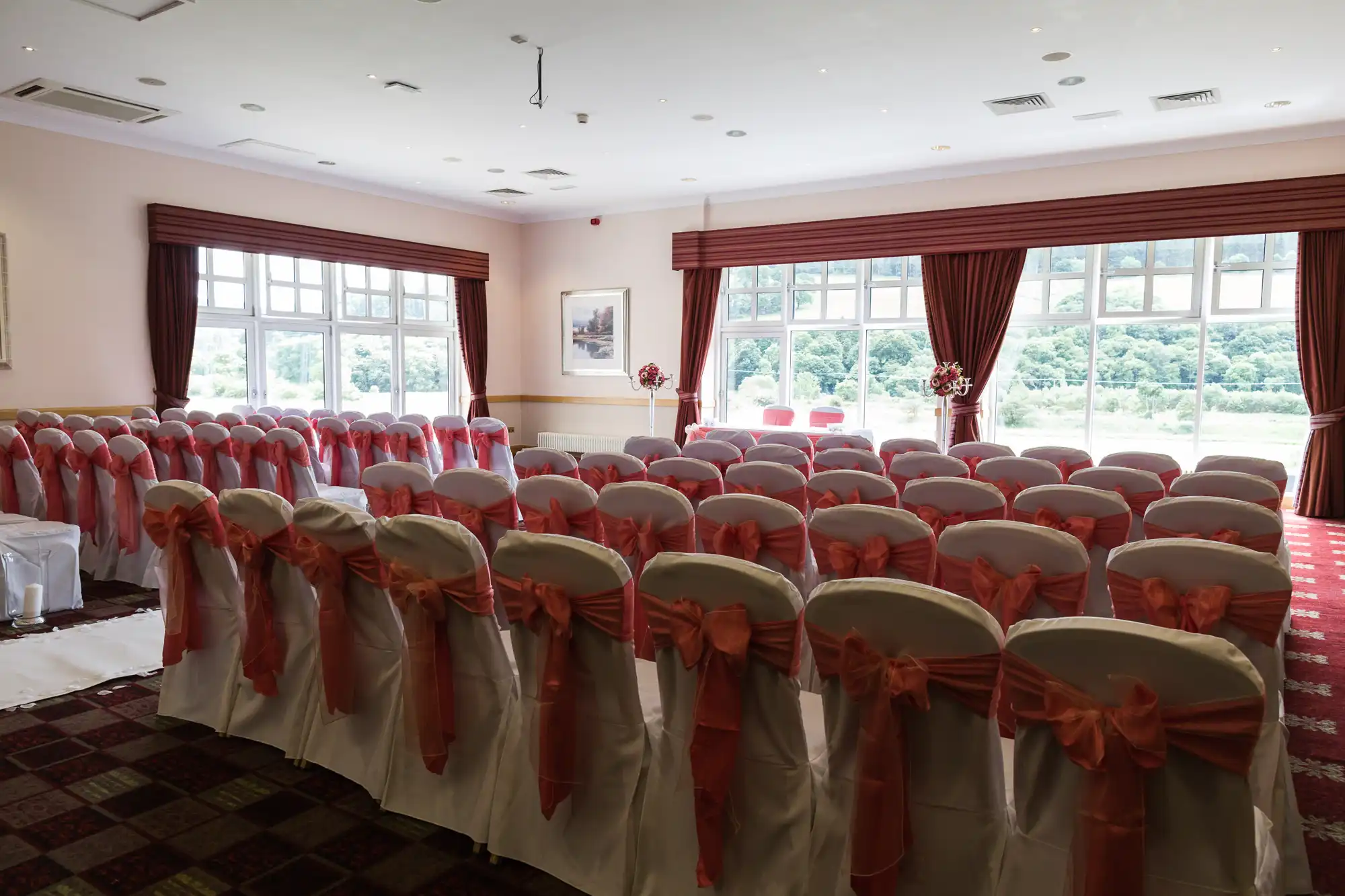 Rows of white chairs with coral sashes are arranged facing forward in a bright room with large windows and red curtains, set up for a wedding ceremony. Two chairs and floral arrangements are visible at the front, with greenery seen outside.