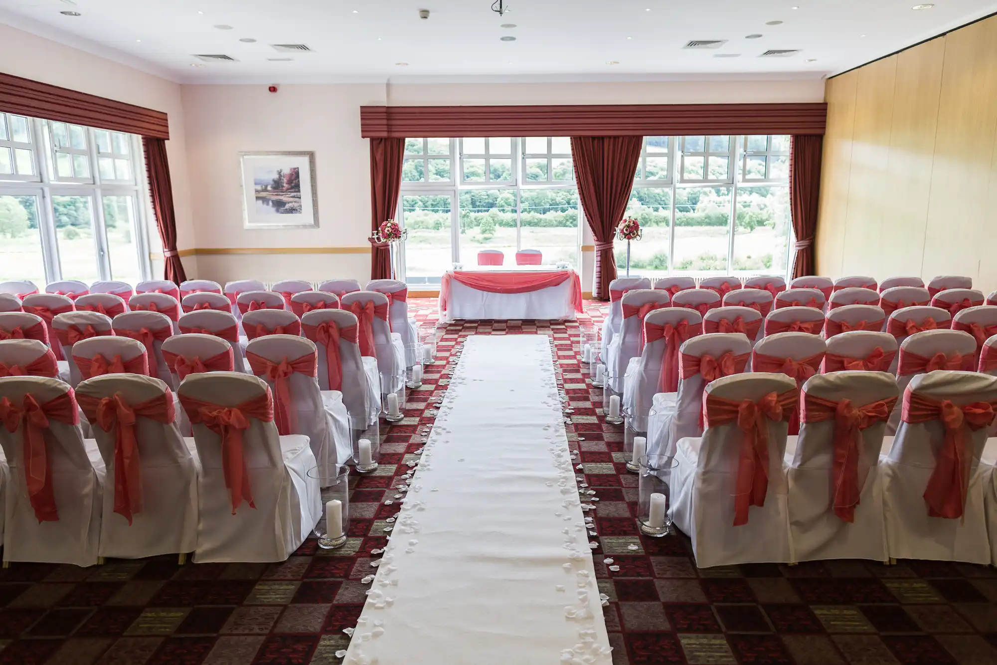 A wedding ceremony room with rows of white chairs adorned with coral bows, facing a table with two chairs at the front. A white aisle runner with flower petals leads to the table, and large windows let in natural light. Red curtains frame the windows.