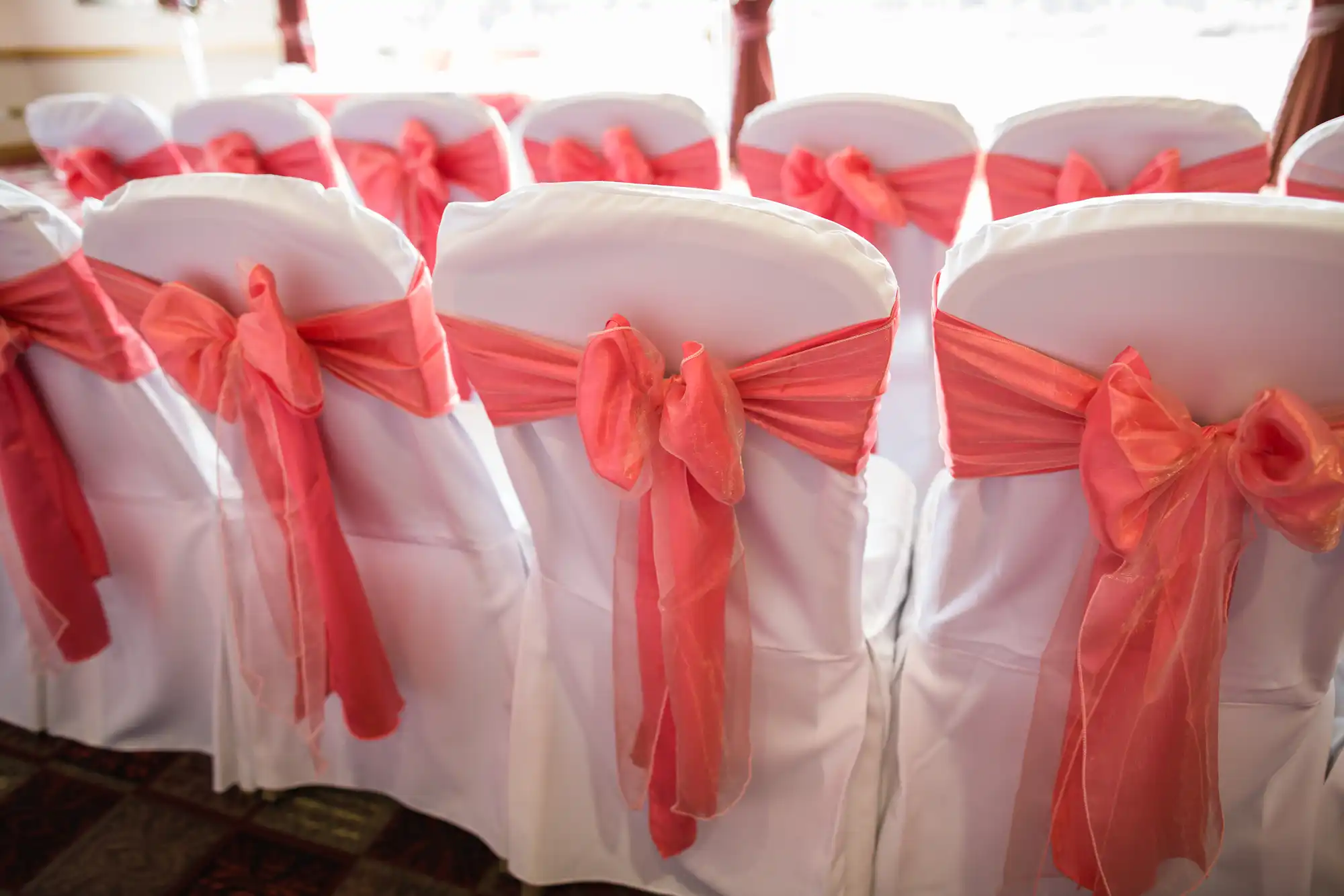 Rows of white chairs are covered with white fabric and decorated with large, sheer coral-pink bows tied around the backs. The chairs are arranged indoors, likely for a formal event such as a wedding or celebration.