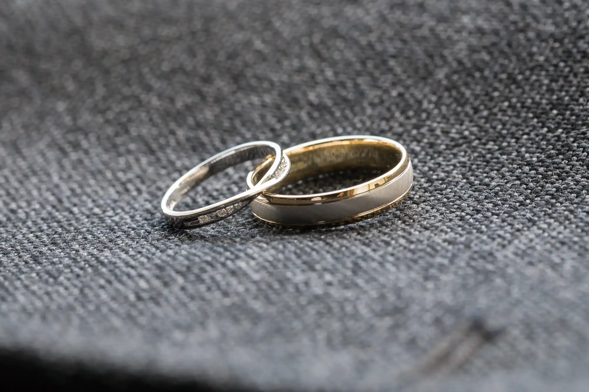 Two rings rest on a textured dark grey fabric. The left ring is thin and silver, while the right ring is wider with a gold edge and a silver centre. Both rings are simple in design, suggesting they may be wedding or engagement bands.