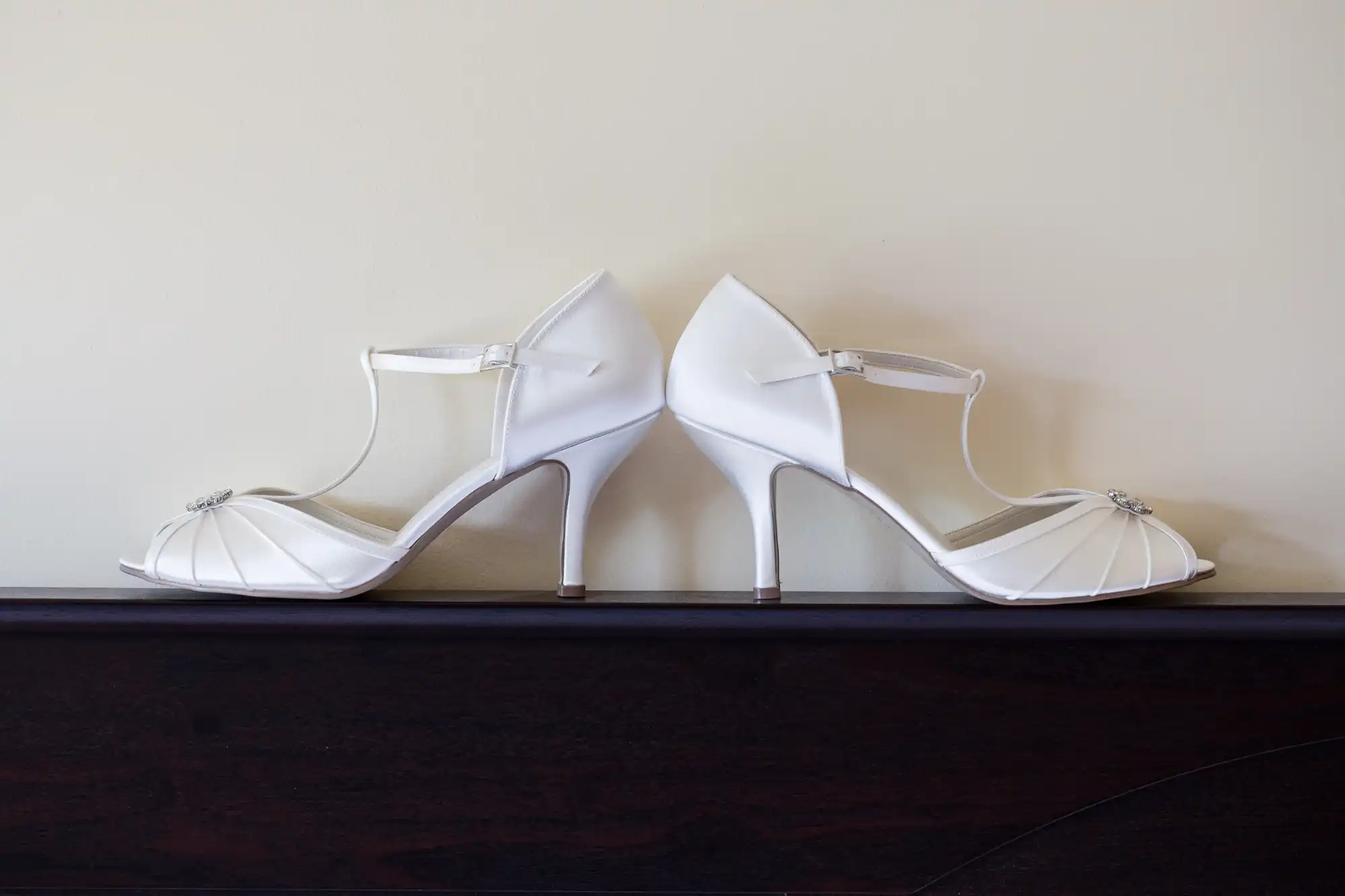 A pair of elegant white high-heeled shoes with ankle straps and delicate pleating detail, embellished with small decorative jewels at the toes, are placed heel to heel on a dark wooden surface against a plain light-coloured wall.