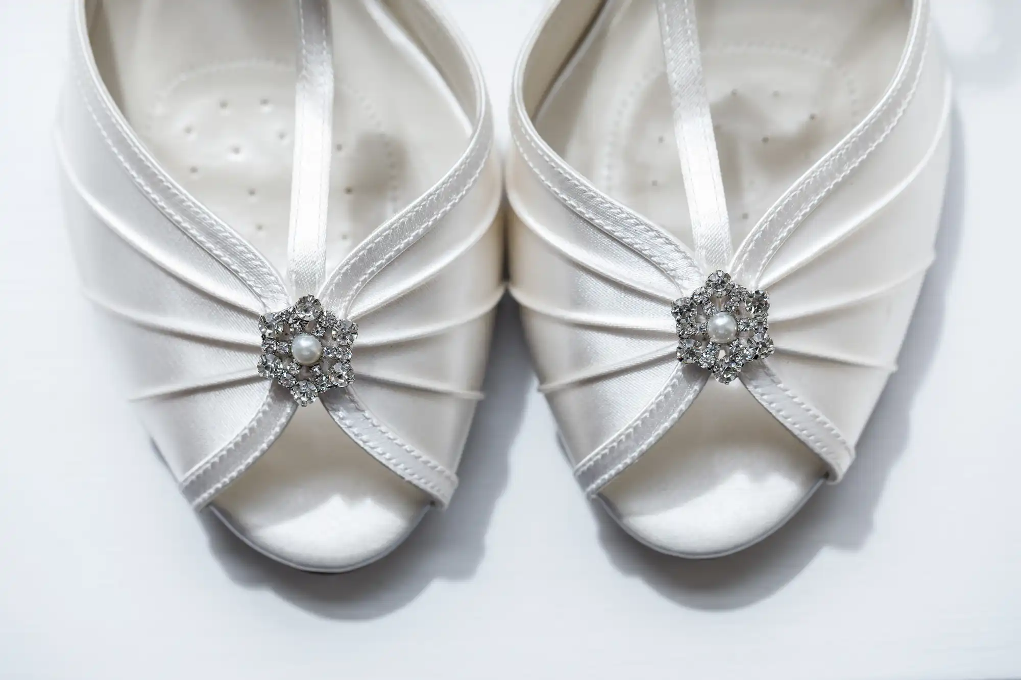 Close-up of a pair of elegant white satin peep-toe shoes with criss-cross straps. Each shoe features a sparkling diamanté and pearl brooch on the front, creating a decorative, bridal look. The background is plain white.