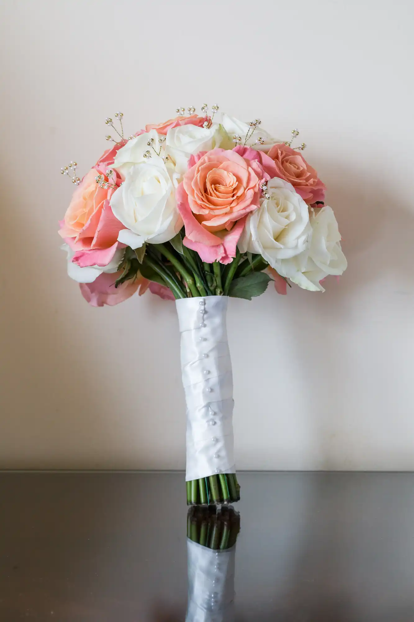 A bouquet of white and peach roses, accented with small white filler flowers, is wrapped with a white satin ribbon. The bouquet stands upright on a reflective surface against a plain, light-coloured background.