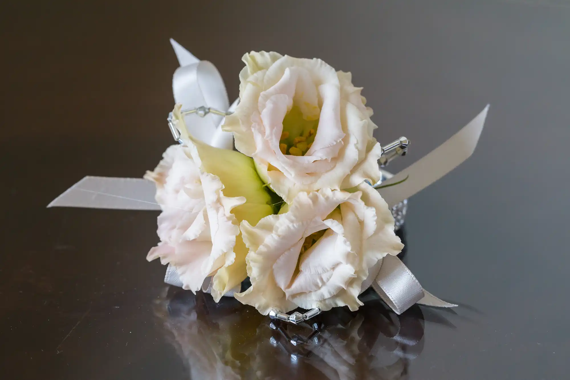 A small corsage featuring three pale cream lisianthus flowers with ruffled petals, accented by green leaves and white ribbon bows, rests on a shiny grey surface. The overall arrangement is neat and elegant.