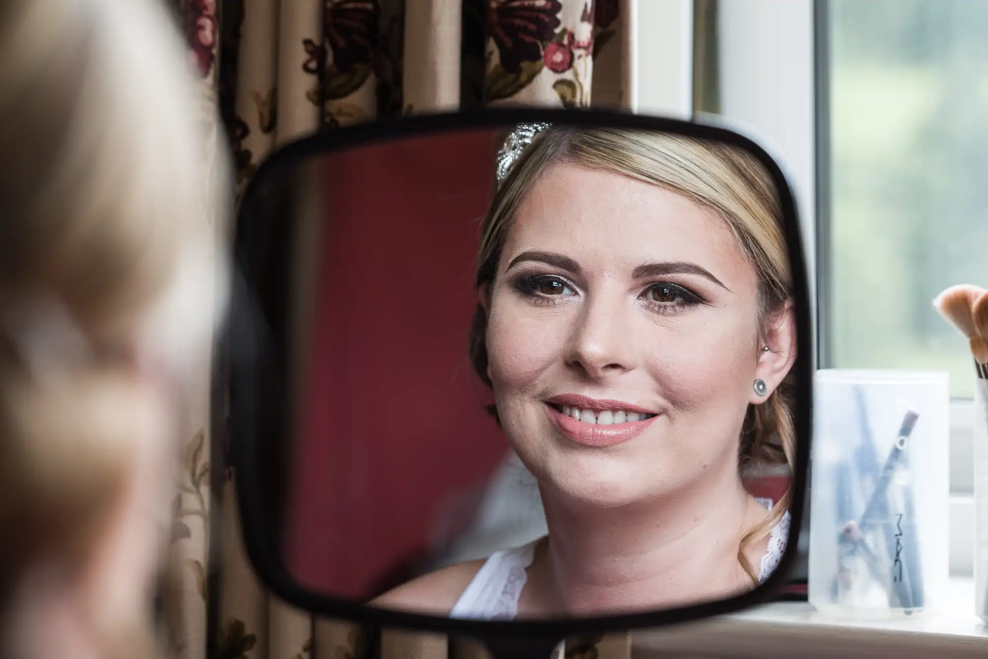 A woman with blonde hair and natural make-up is smiling as she looks at her reflection in a rectangular mirror. The background shows a window, floral curtains, and a clear container holding make-up brushes.