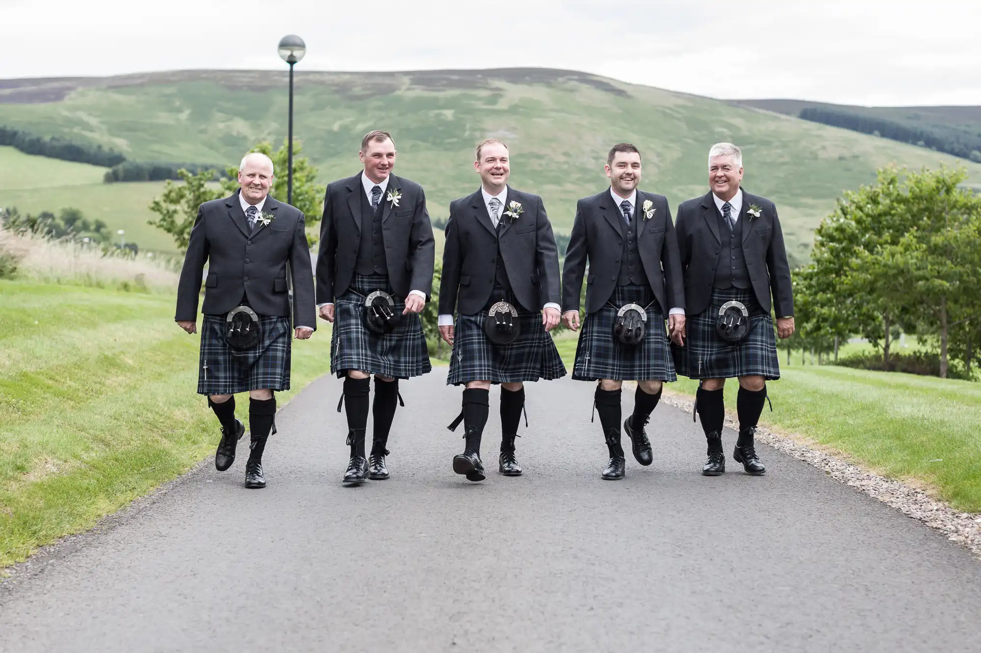 Five men in black jackets, kilts, and knee-high socks walk together and smile outdoors on a paved path, with green hills, grass, trees, and cloudy sky in the background. They appear to be dressed for a formal or celebratory occasion.