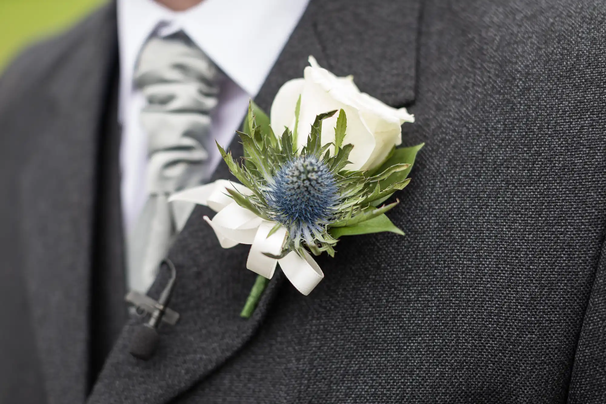 Close-up of a man’s suit jacket with a buttonhole pinned on the lapel. The buttonhole features a white rose, a blue thistle flower, greenery, and a white ribbon. The man is also wearing a silver patterned tie and a white dress shirt.