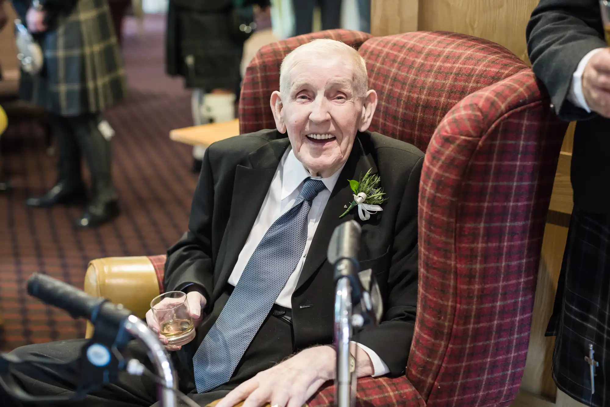 An elderly man in a suit and tie sits smiling in a red checked armchair, holding a drink in one hand. He wears a buttonhole on his lapel. A walking frame is in front of him and other people in formal attire are visible in the blurred background.