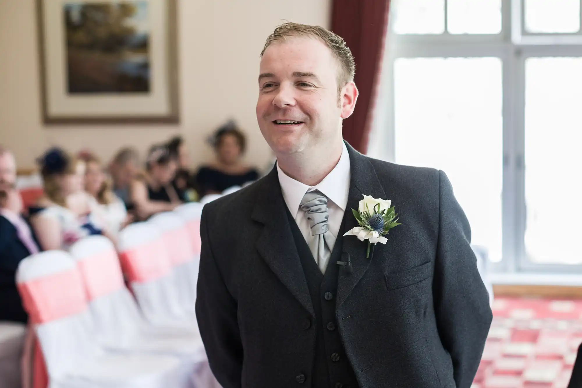 A smiling man in a grey suit with a silver tie and flower buttonhole stands indoors at an event, likely a wedding. Behind him, people are seated on chairs with white and pink covers. Bright windows and a framed picture are in the background.