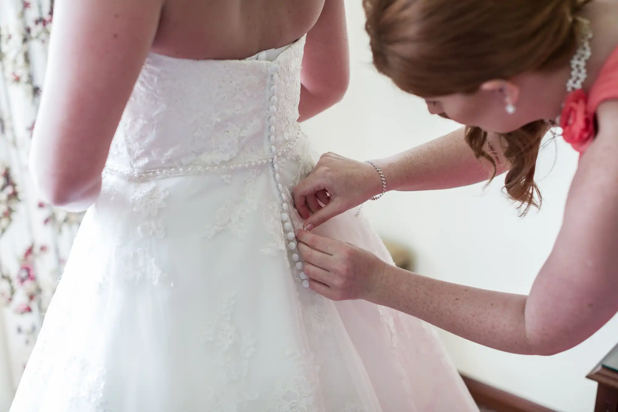 A woman in a coral dress helps button the back of a bride’s white lace wedding gown. The bride’s dress features floral lace details, a row of buttons, and a beaded belt. The scene is indoors with soft lighting and floral curtains partly visible.