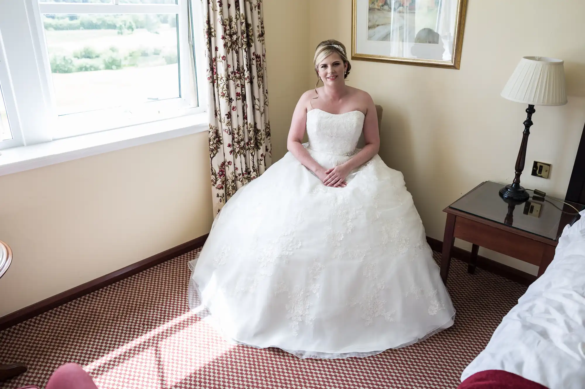 A bride in a white strapless wedding dress sits on the edge of a bed in a softly lit hotel room, hands in her lap, smiling. The room features cream walls, a window with floral curtains, a framed picture, lamp, bedside table, and red carpeted floor.