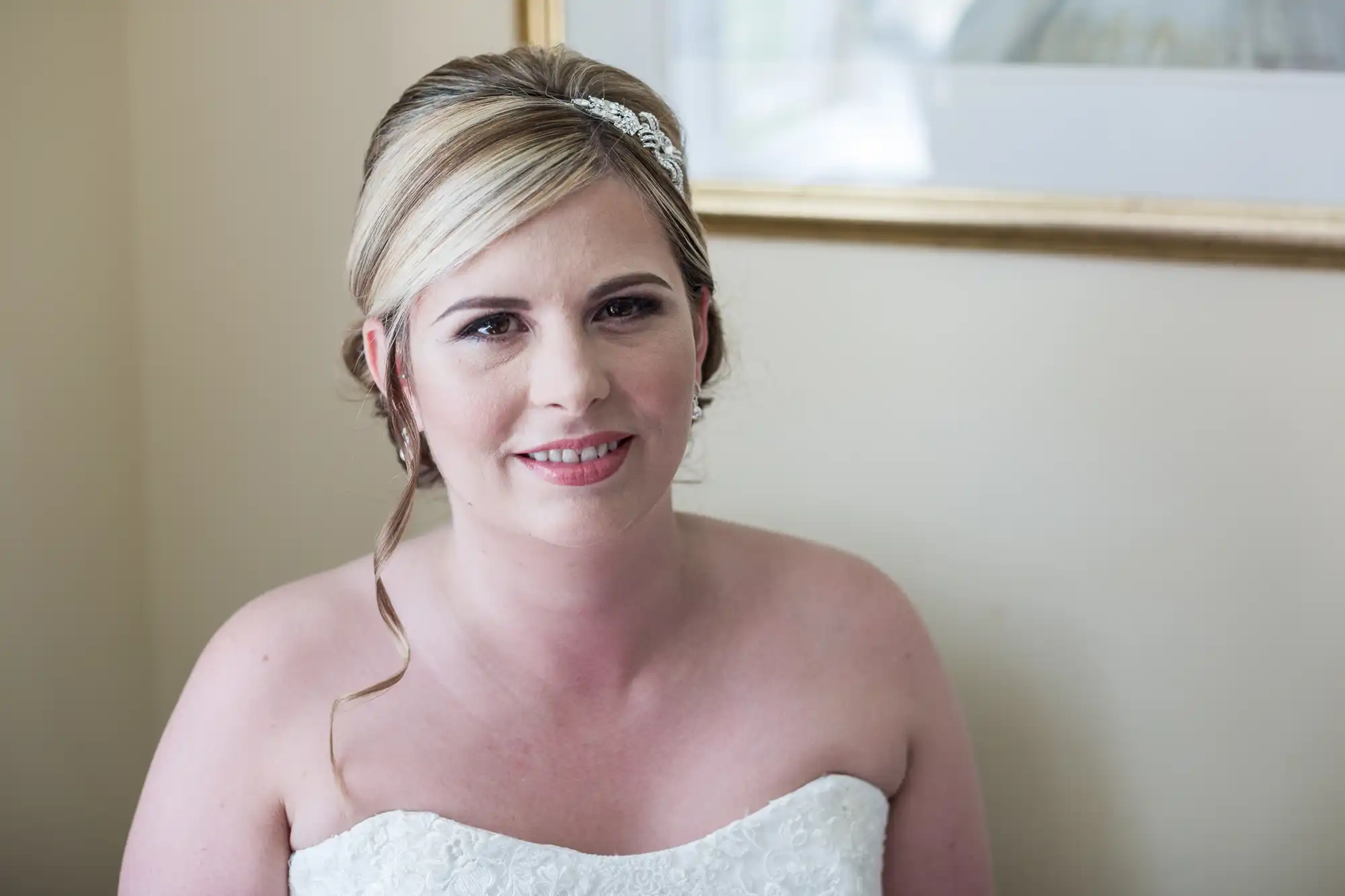 A woman in a white strapless wedding dress sits indoors, smiling gently. She has fair skin, blonde hair styled up with a jewelled headband, and natural make-up. A framed picture hangs on the wall behind her. The lighting is soft and natural.