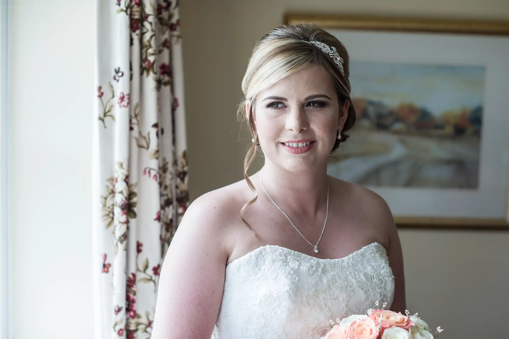 A smiling bride in a white lace strapless wedding dress stands indoors near a window with floral curtains, holding a bouquet of pink and white flowers. She has fair hair styled up with a jewelled headband and wears a delicate necklace.