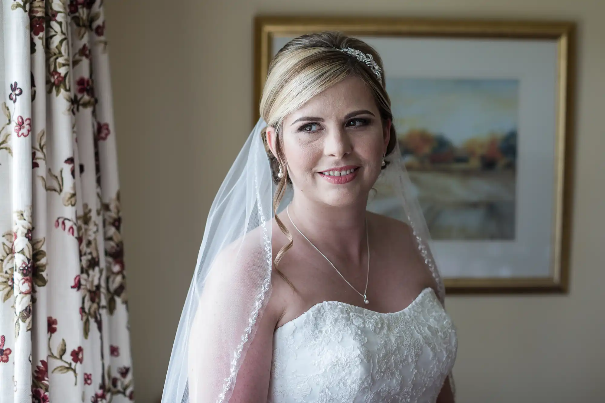 A woman in a white strapless wedding dress and veil stands indoors, smiling softly. She has fair hair styled up with a jewelled headband. Floral curtains and a framed landscape painting are visible in the background.