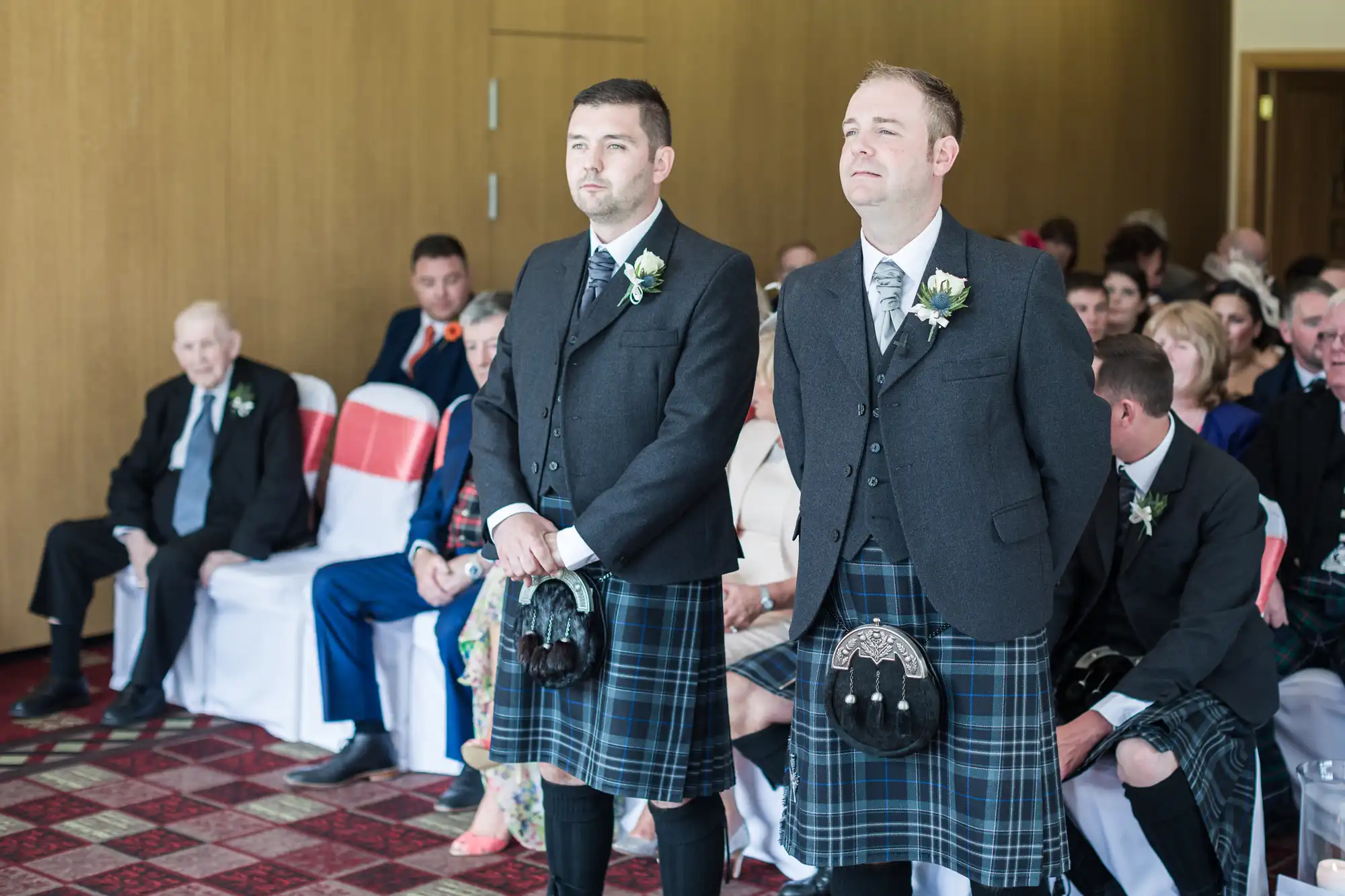 Two men wearing black jackets, white shirts, ties, and traditional Scottish kilts stand at the front of a room. They have buttonholes on their lapels. Several seated guests, some elderly, watch from chairs, suggesting a formal event, likely a wedding.
