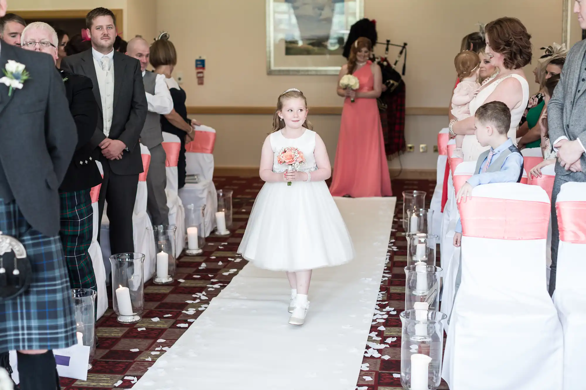 A young girl in a white dress walks down an aisle lined with flower petals and candles, holding a bouquet. She’s surrounded by seated and standing wedding guests, some in formal attire. White chair covers with pink sashes decorate the chairs.