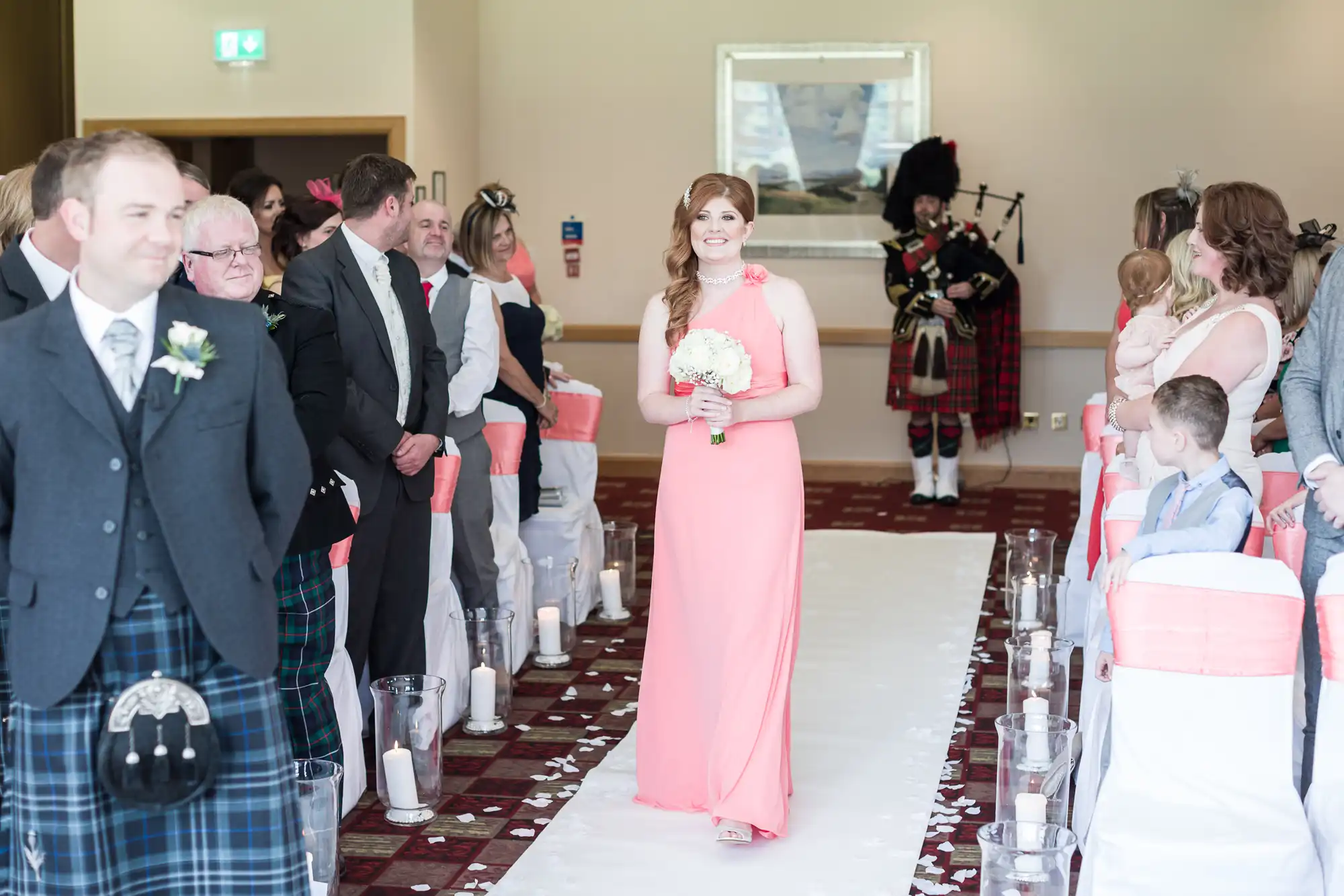 A woman in a long pink dress holds a bouquet as she walks down an aisle lined with white and pink chairs and candles. Guests watch her, some smiling. A man in a kilt stands at the front; a bagpiper in Scottish attire plays in the background.