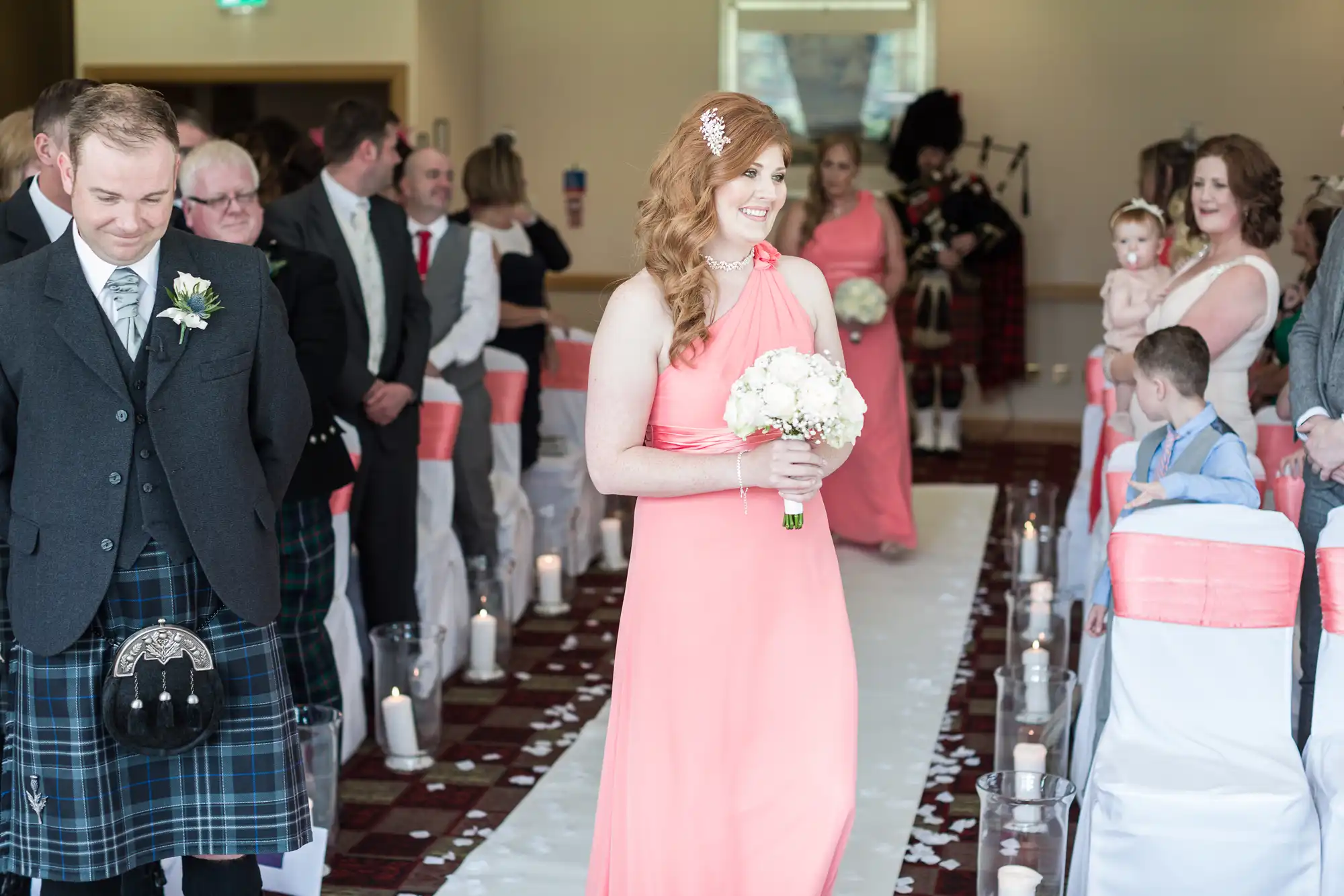 A smiling bridesmaid in a peach dress holds a bouquet whilst walking down an aisle lined with guests. Men in kilts, candles on the floor, and rose petals decorate the aisle. People watch, some smiling; a woman holds a baby. It is an indoor wedding ceremony.