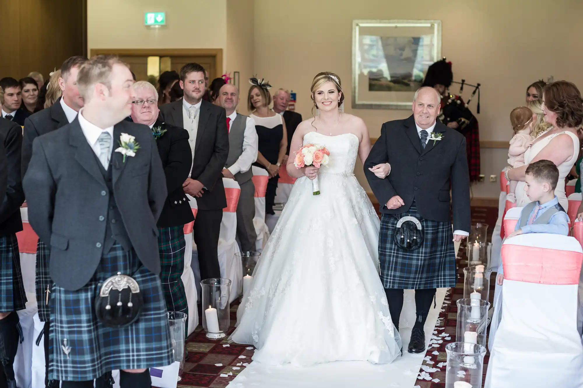 A bride in a white gown holding peach flowers walks down the aisle, arm-in-arm with an older man in a kilt. Guests, including children, watch. Men wear kilts and jackets; women are in dresses. The aisle is lined with candles, and the setting is a bright indoor venue.