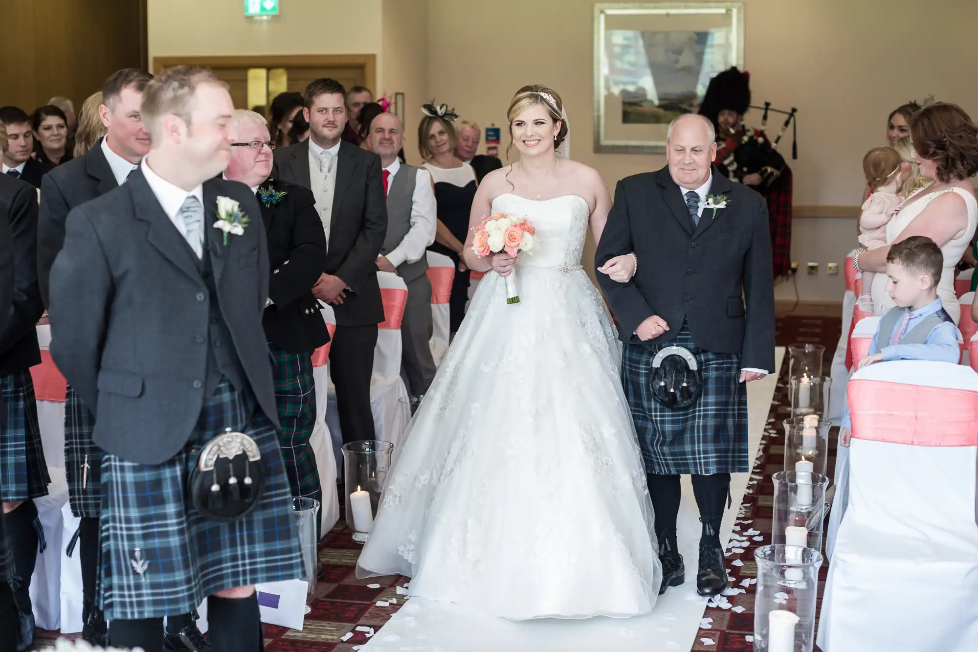 A smiling bride in a white gown holds a pastel bouquet and walks down the aisle with an older man in a kilt. Guests in formal attire, some in kilts, watch and smile. The aisle is lined with white petals and candles; chairs have pink sashes.