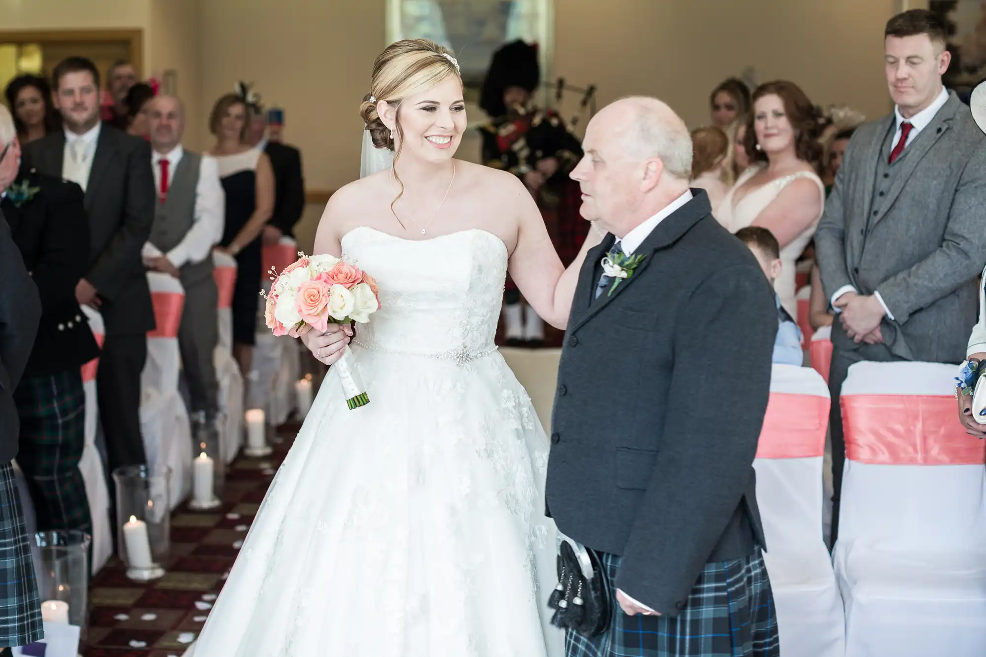 A bride in a white dress holds a bouquet and smiles at an older man, possibly her father, as they stand in an aisle with seated guests watching. The guests are dressed formally, and the chairs have white covers with pink sashes. Candles line the aisle.