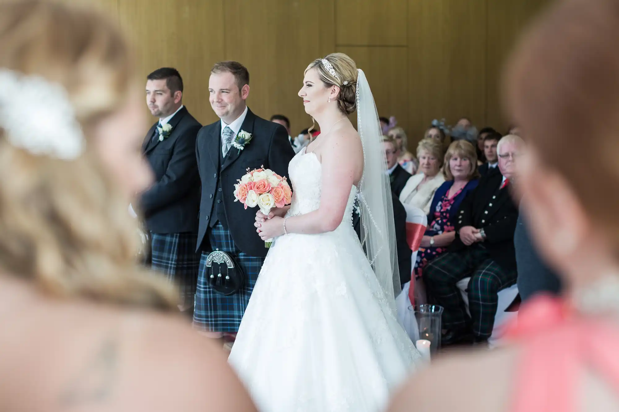 A bride in a white gown and veil holds a bouquet of peach and white flowers, standing beside two men in kilts. They face a seated audience in a bright room with wooden walls. Foreground shows blurred guests, focussing attention on the wedding ceremony.