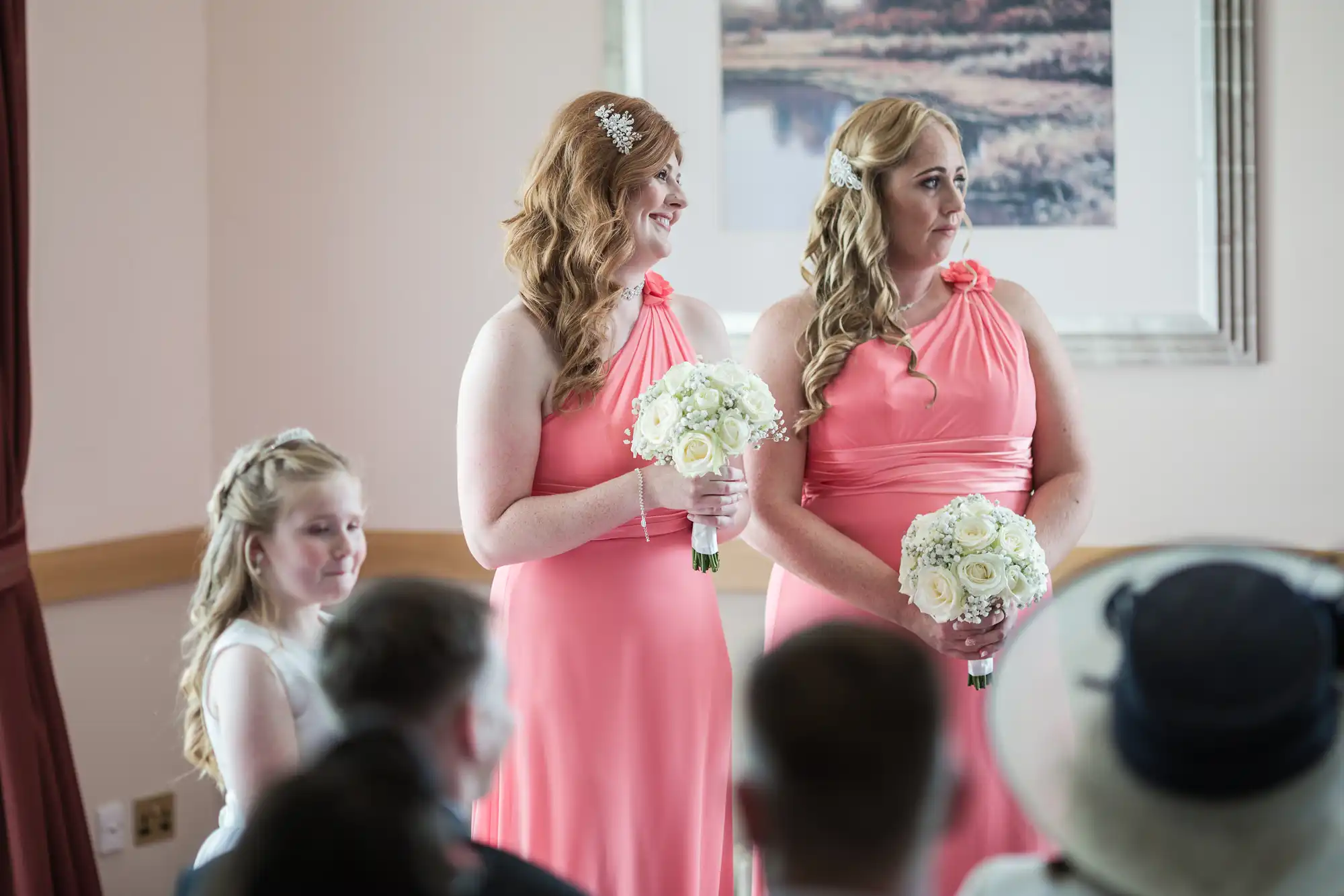 Two women in matching coral pink dresses hold white flower bouquets and smile, standing side by side. A young girl in white sits nearby. All are indoors, with seated guests visible in the foreground and a framed picture on the wall behind them.