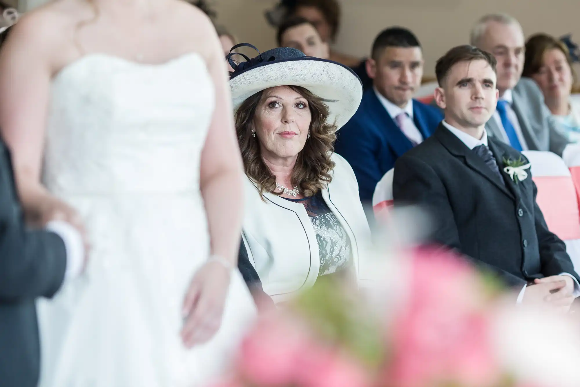 A woman in a large hat and formal attire sits among guests at a wedding ceremony, looking thoughtfully ahead. The blurred bride in a white dress stands beside her, while other seated guests in suits and dresses watch attentively in the background.