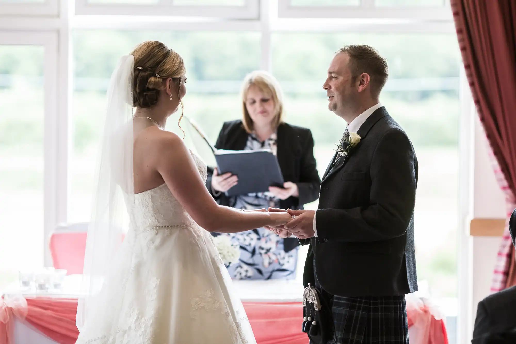 A bride in a white dress and veil holds hands with a groom in a dark jacket and kilt during a wedding ceremony, while an officiant reads from a book in the background. Sunlight streams through large windows behind them.