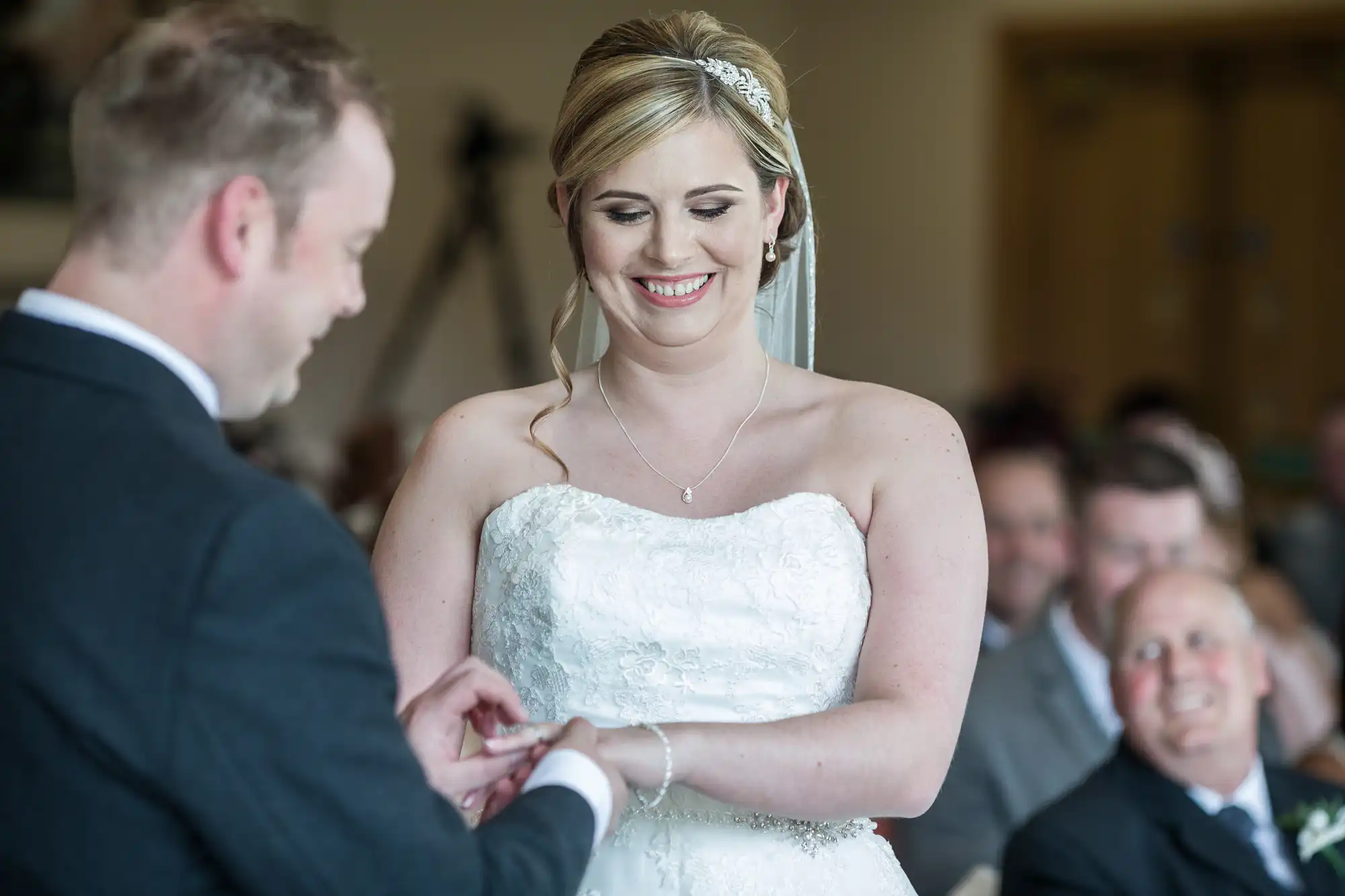 A bride in a white lace strapless gown smiles as a groom in a dark suit places a ring on her finger during a wedding ceremony. Guests watch and smile in the blurred background. The moment is joyful and intimate.