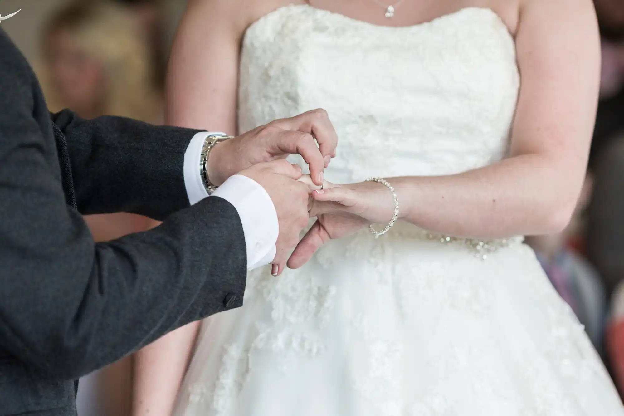 A close-up of a bride in a white lace wedding dress as her partner, in a dark suit, gently places a ring on her finger during a wedding ceremony. The focus is on their hands and the ring exchange, with soft light and blurred background.