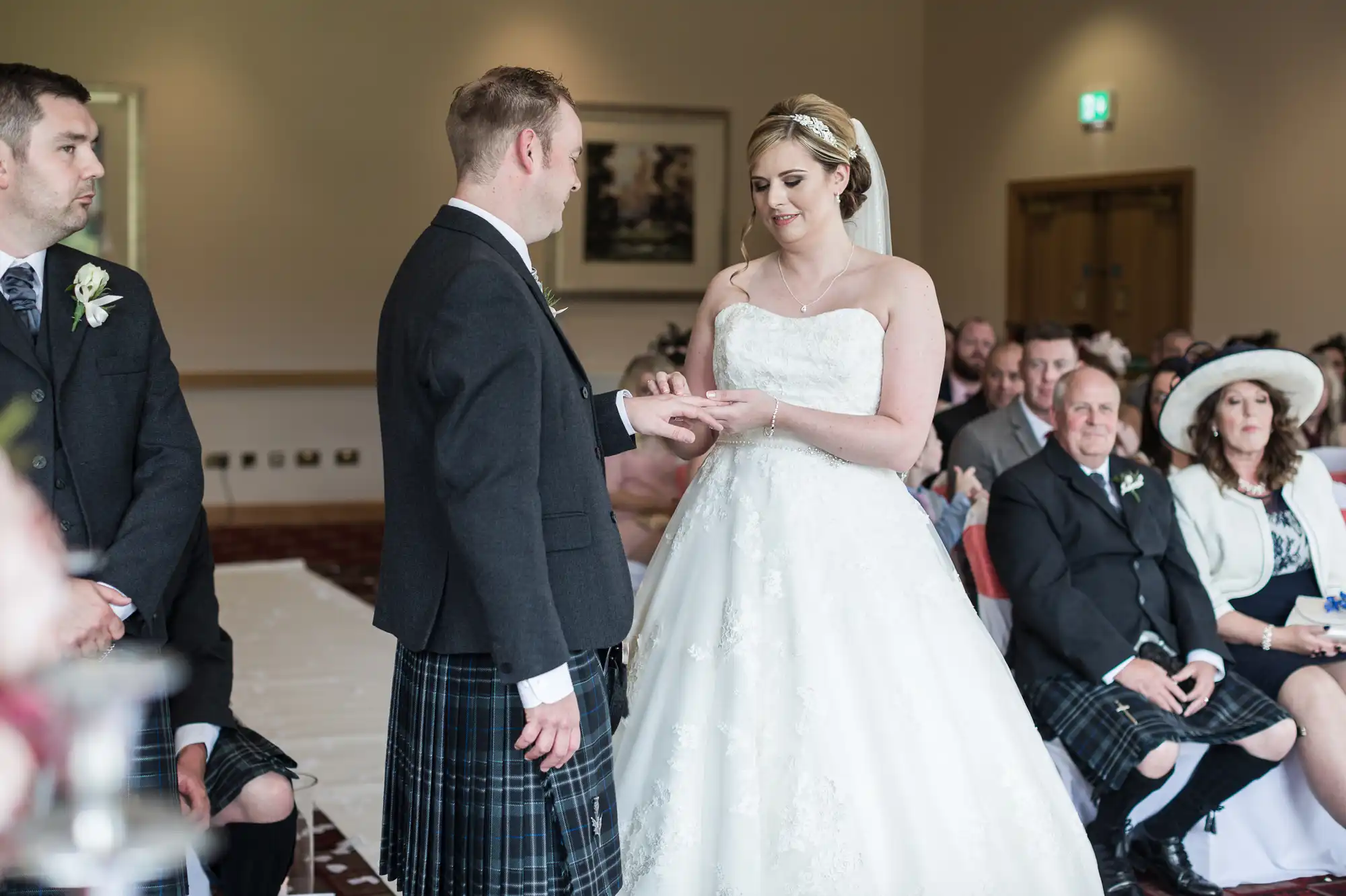 A groom in a black jacket and tartan kilt places a ring on the bride's finger during their wedding ceremony. The bride wears a white strapless dress and tiara. Guests, some in kilts, watch from their seats in a light-filled room.