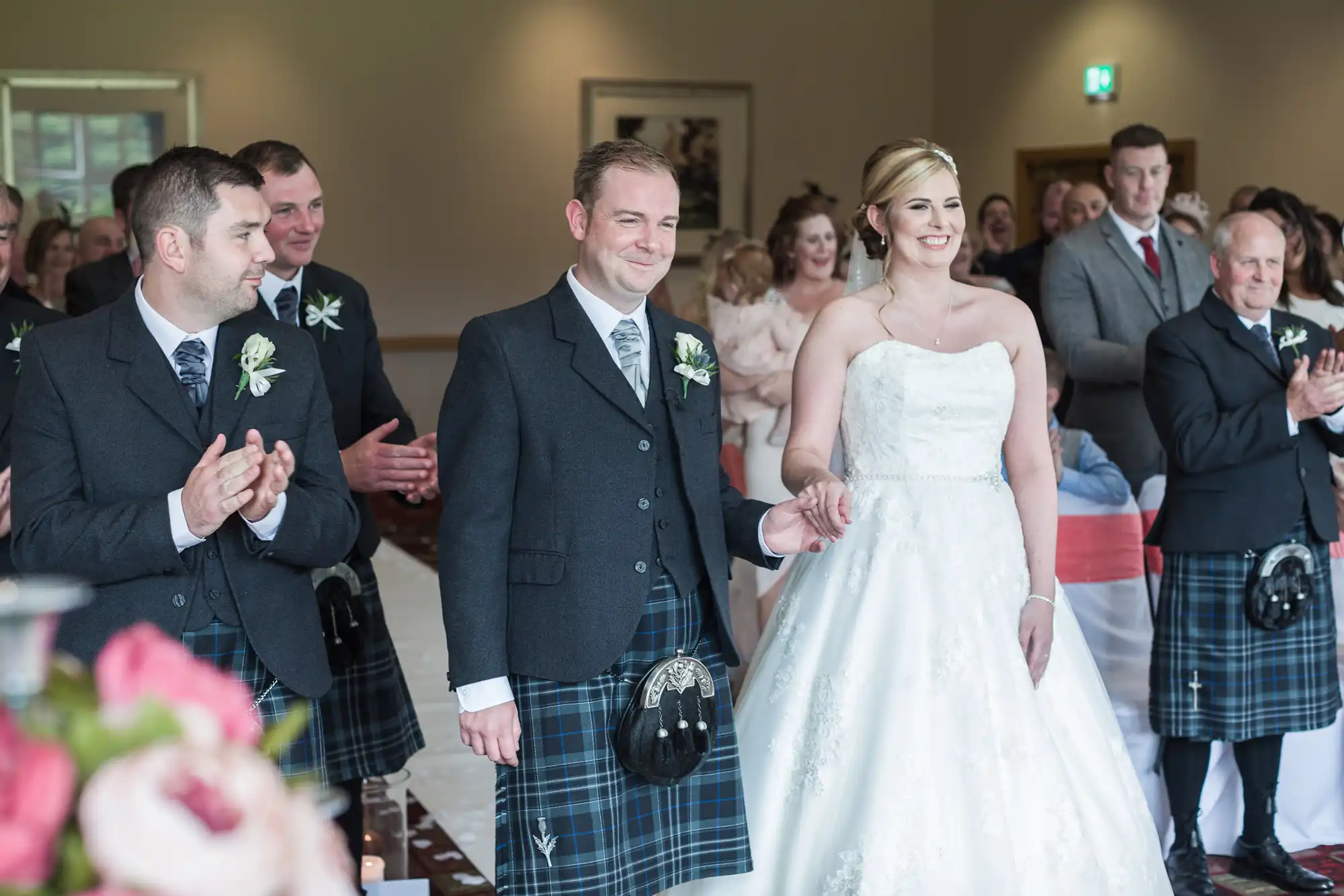 A bride in a white dress and a groom in a black jacket and tartan kilt stand smiling and holding hands at the aisle, surrounded by applauding guests. The indoor venue is decorated in soft colours, with flower arrangements visible in the foreground.