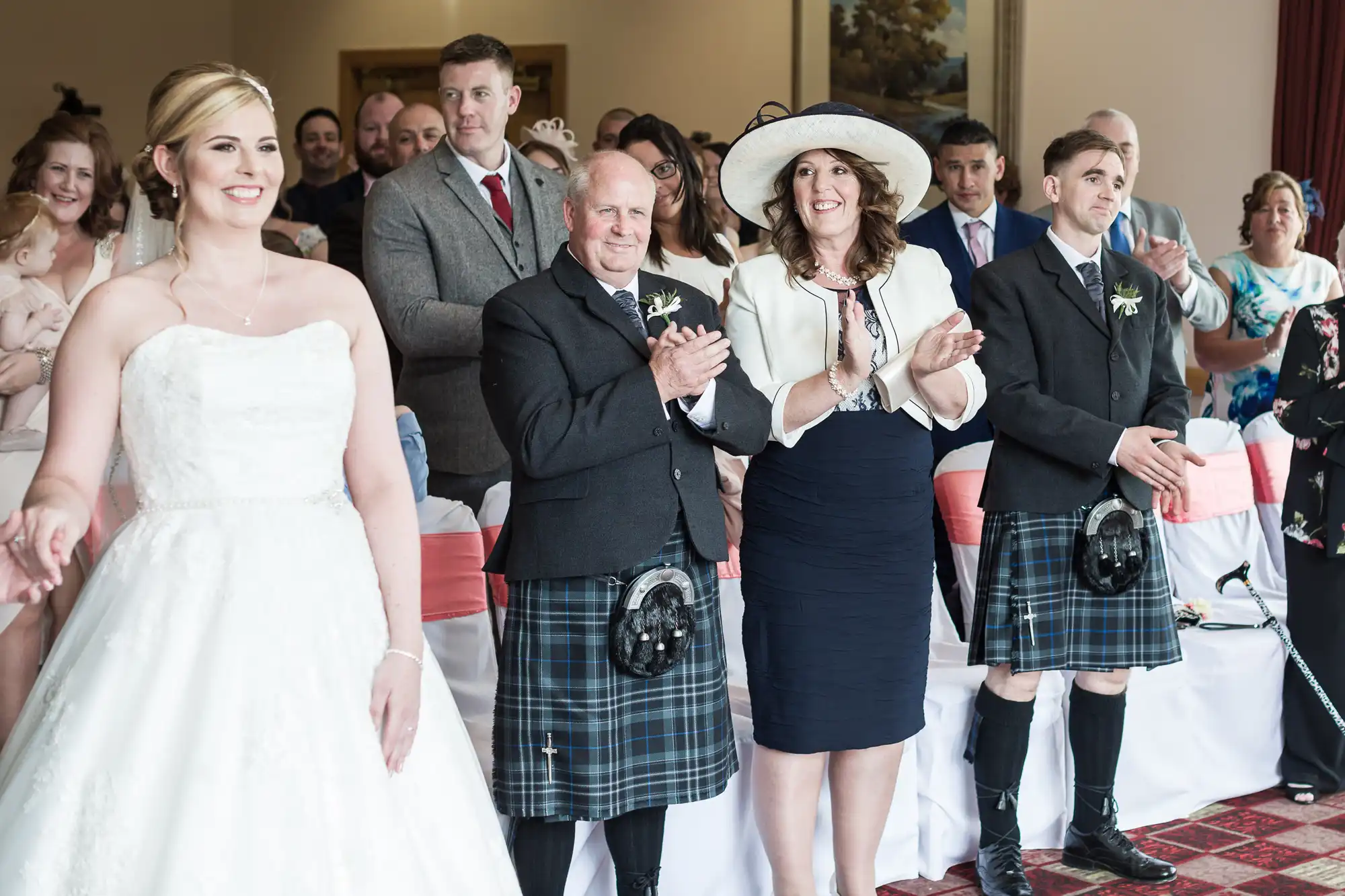 A smiling bride in a white dress stands in front of seated and standing guests, including two men in kilts and a woman in a white hat and jacket, all applauding and smiling in a bright room decorated for a wedding ceremony.