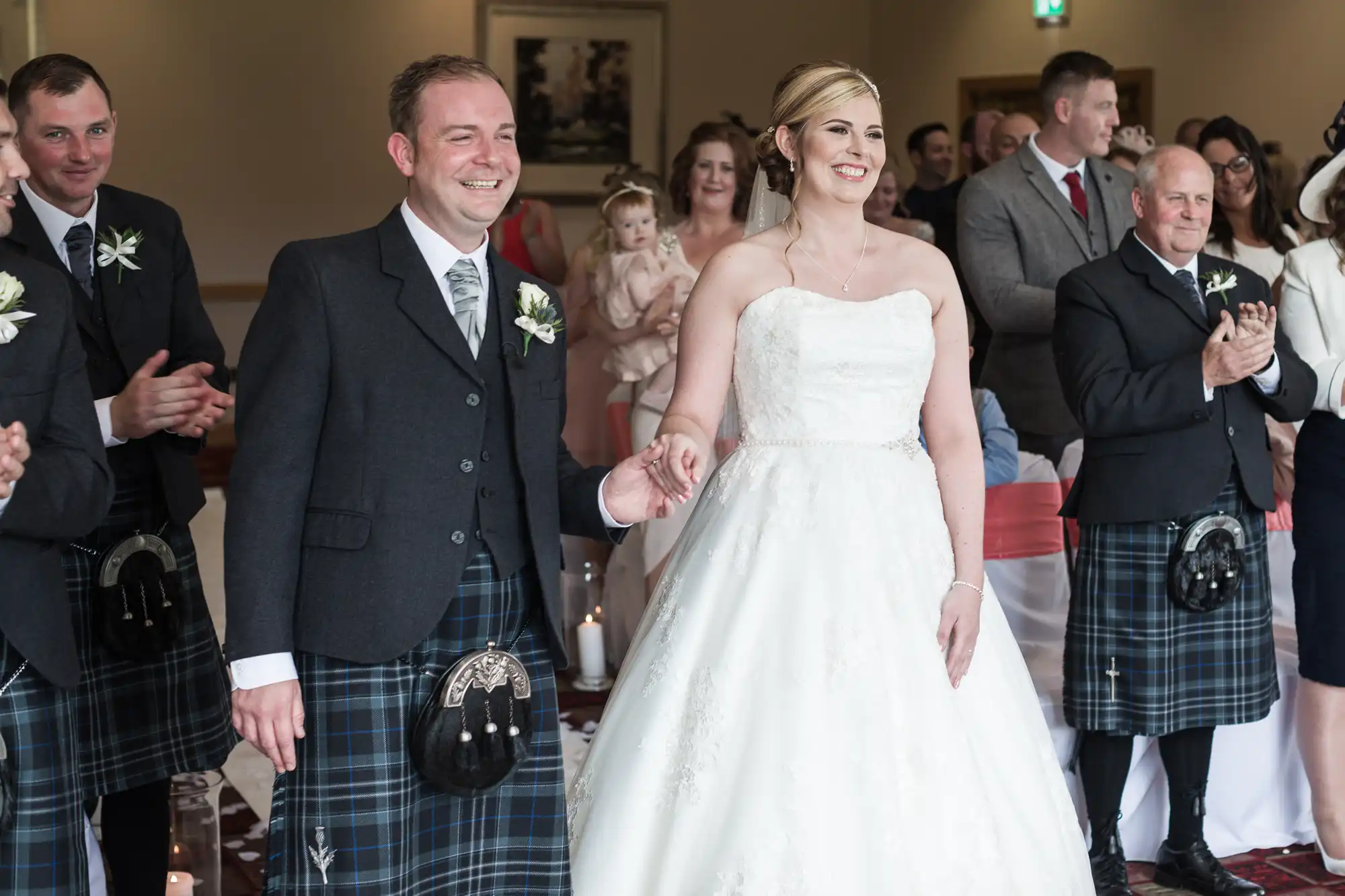 A bride in a white strapless gown and a groom in a black jacket and tartan kilt hold hands, smiling, as they walk down the aisle. Guests, many in kilts, stand and clap. A woman holds a baby in the background. The setting is a bright, elegant room.
