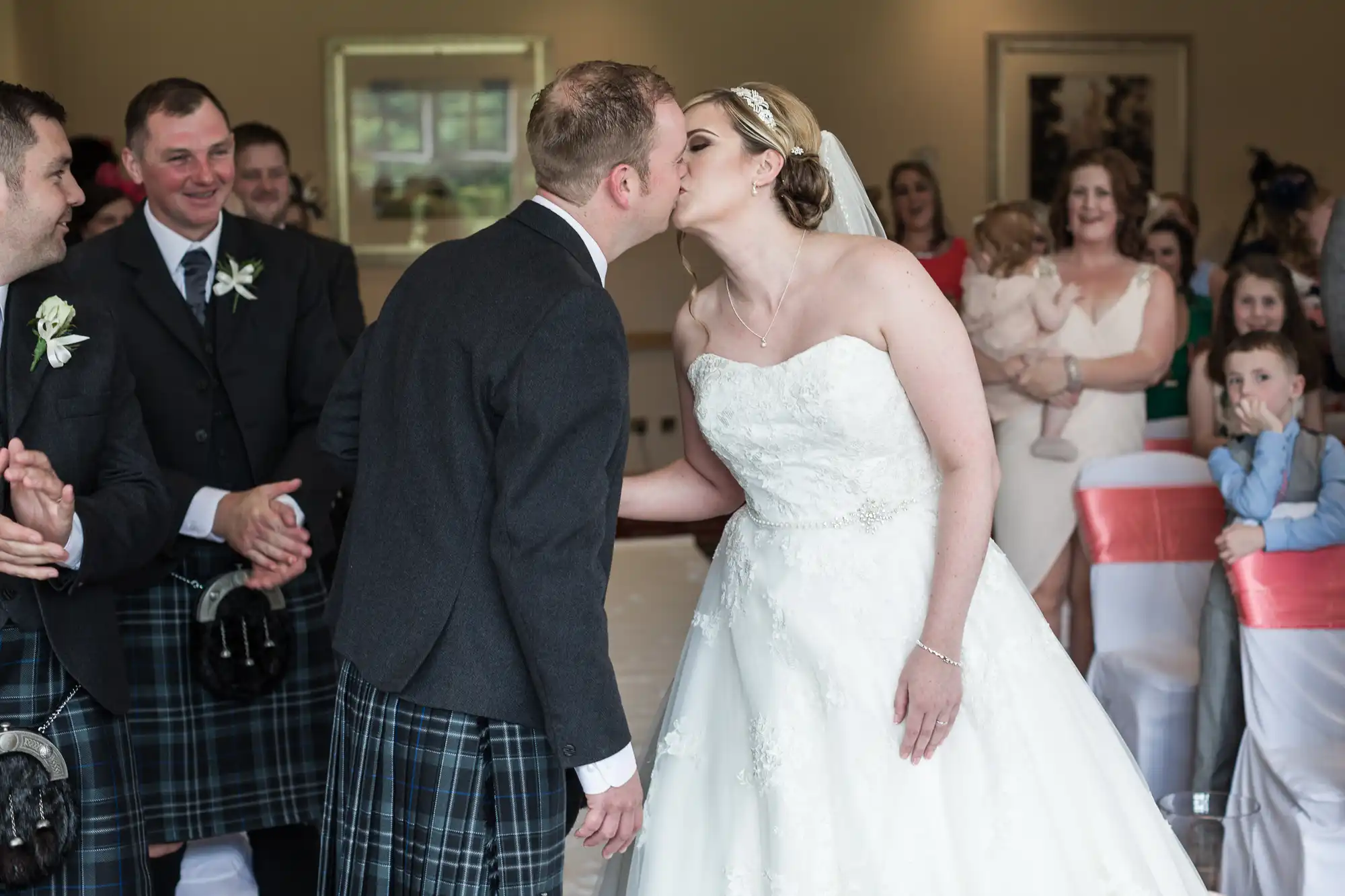 A bride in a white strapless gown and a groom in a dark jacket and tartan kilt kiss at the aisle, surrounded by guests in kilts and formal attire, some clapping and smiling. Chairs have red and white covers, and children watch in the background.