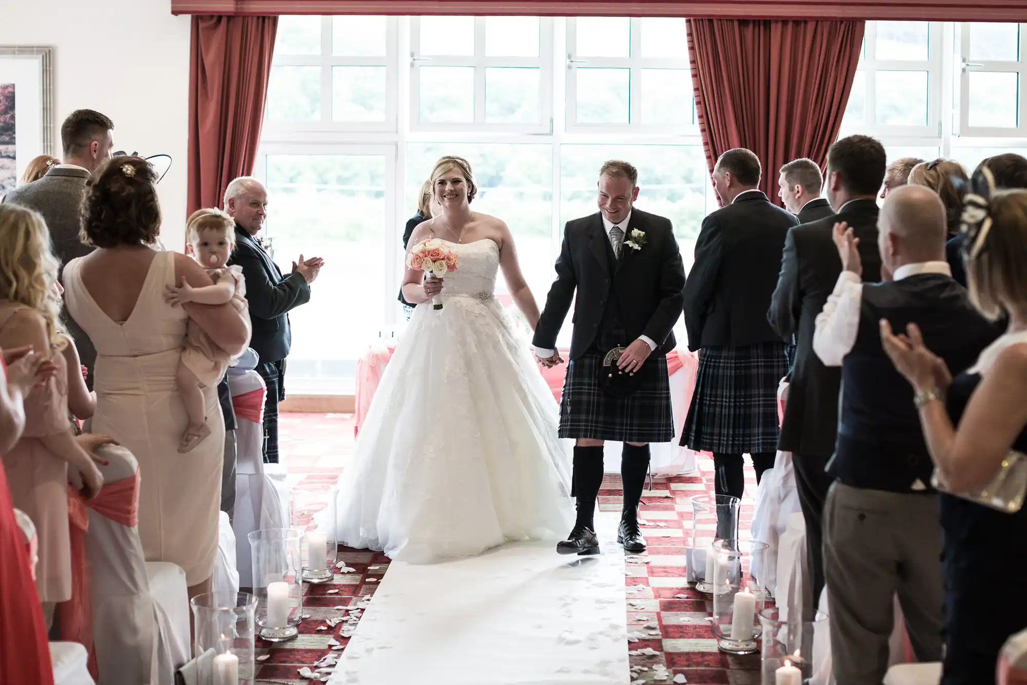A bride in a white gown holding pink flowers walks down the aisle with a man in a black jacket and kilt. Guests, some applauding and one holding a child, stand on either side. The room has red curtains, white chairs, and candles along the aisle.