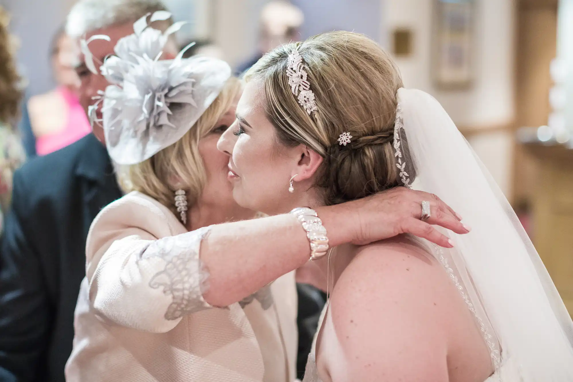 A bride in a white dress and veil hugs an older woman wearing a light-coloured hat and jacket. The older woman’s face is partly hidden as she embraces the bride, who smiles with closed eyes. Other people are blurred in the background.