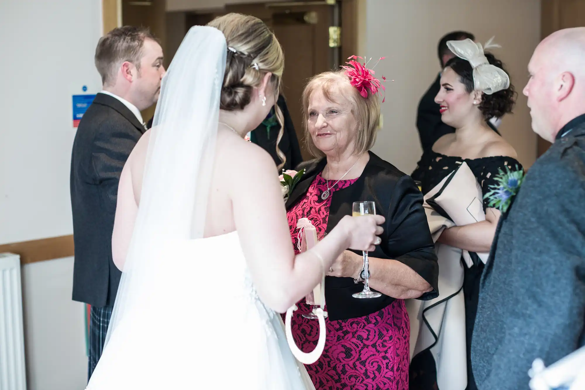 A bride in a white dress and veil holds a champagne glass, speaking to an older woman in a pink and black dress with a pink fascinator. Other guests, dressed formally, stand nearby, chatting at an indoor wedding reception.